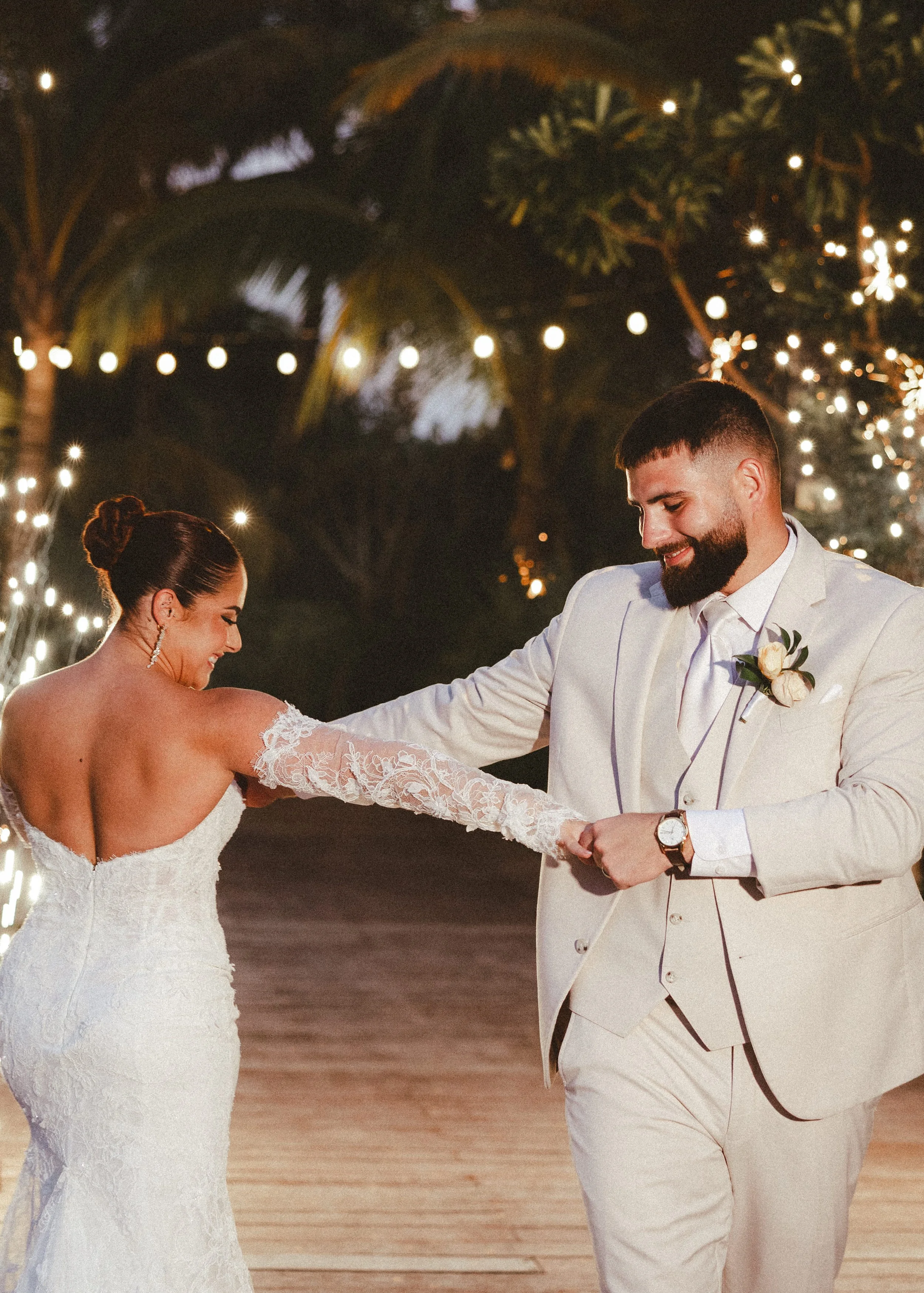 A bride and groom are dancing during their outdoor wedding reception at night, decorated with string lights and tropical trees.