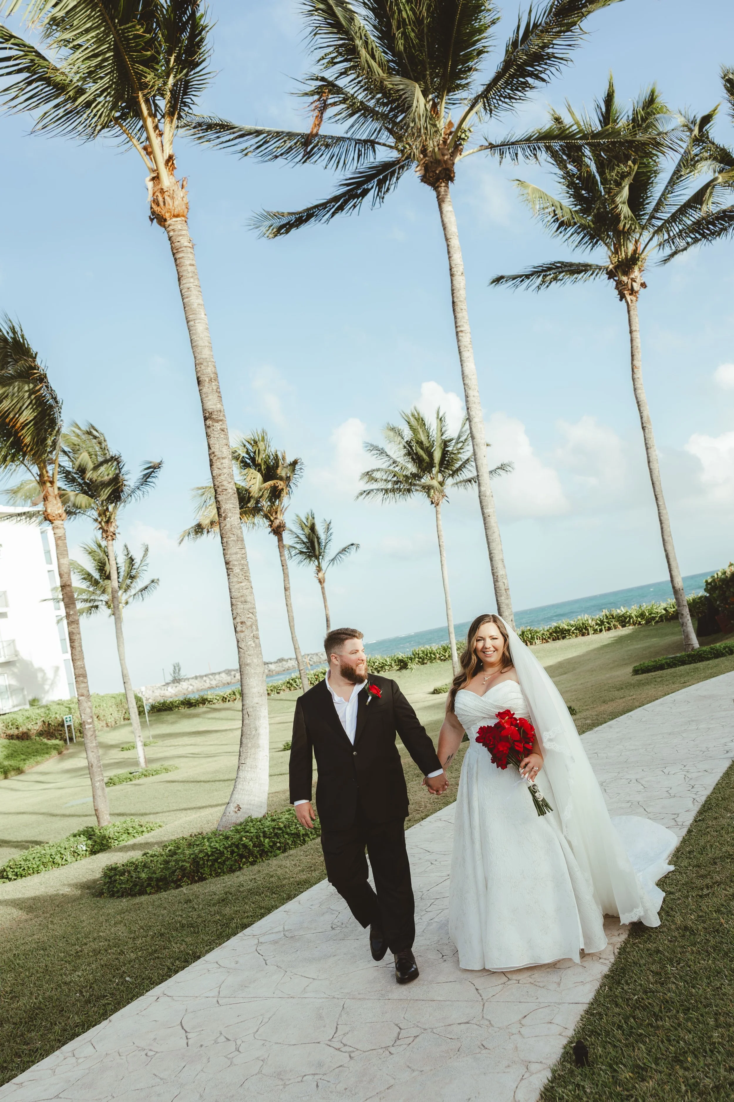 A bride and groom walking hand in hand outdoors on a sunny day, with palm trees, grass, and the ocean in the background in Dreams Jade.