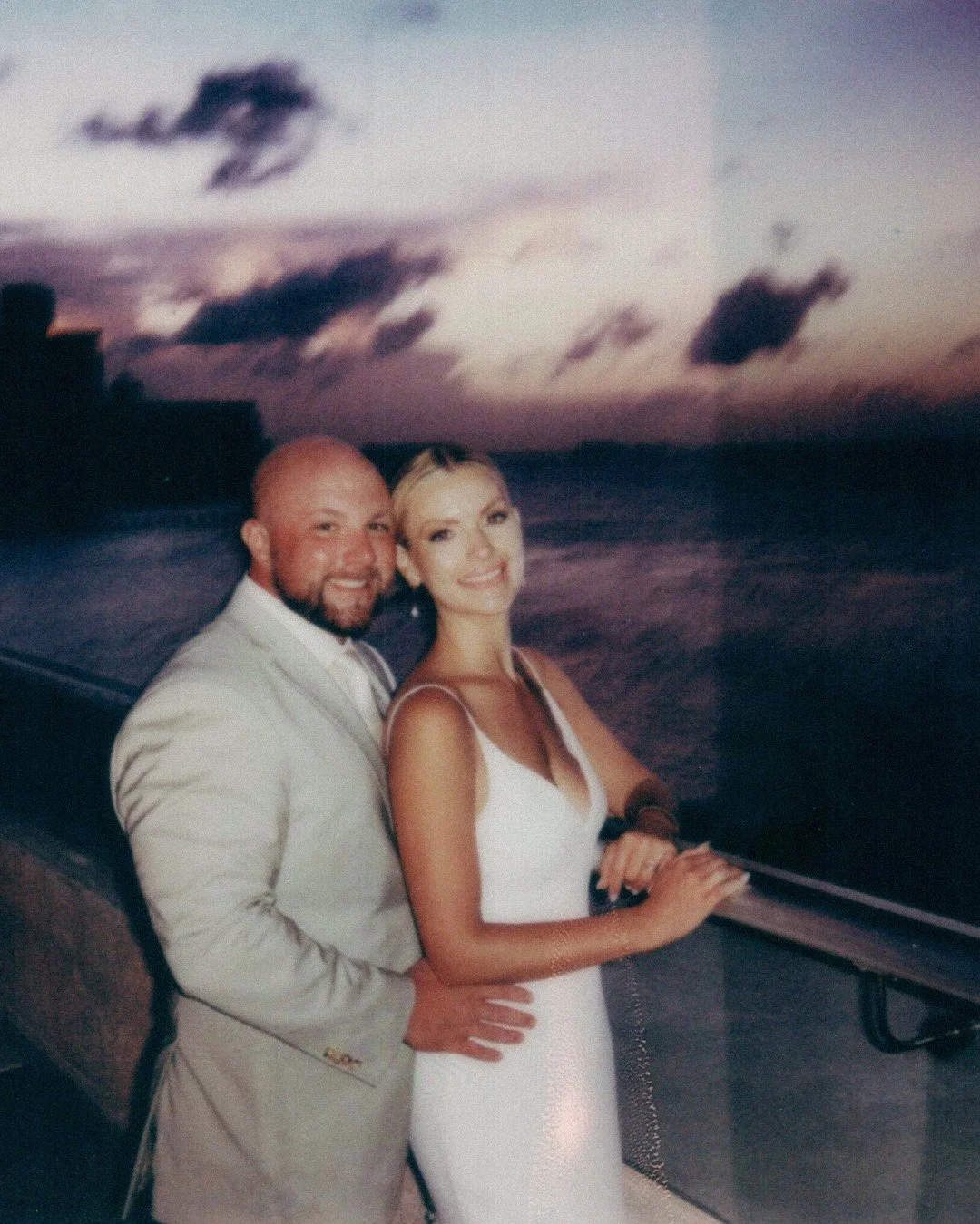A couple dressed in wedding attire standing on a boat at sunset, with a dark ocean and cloudy sky in the background.