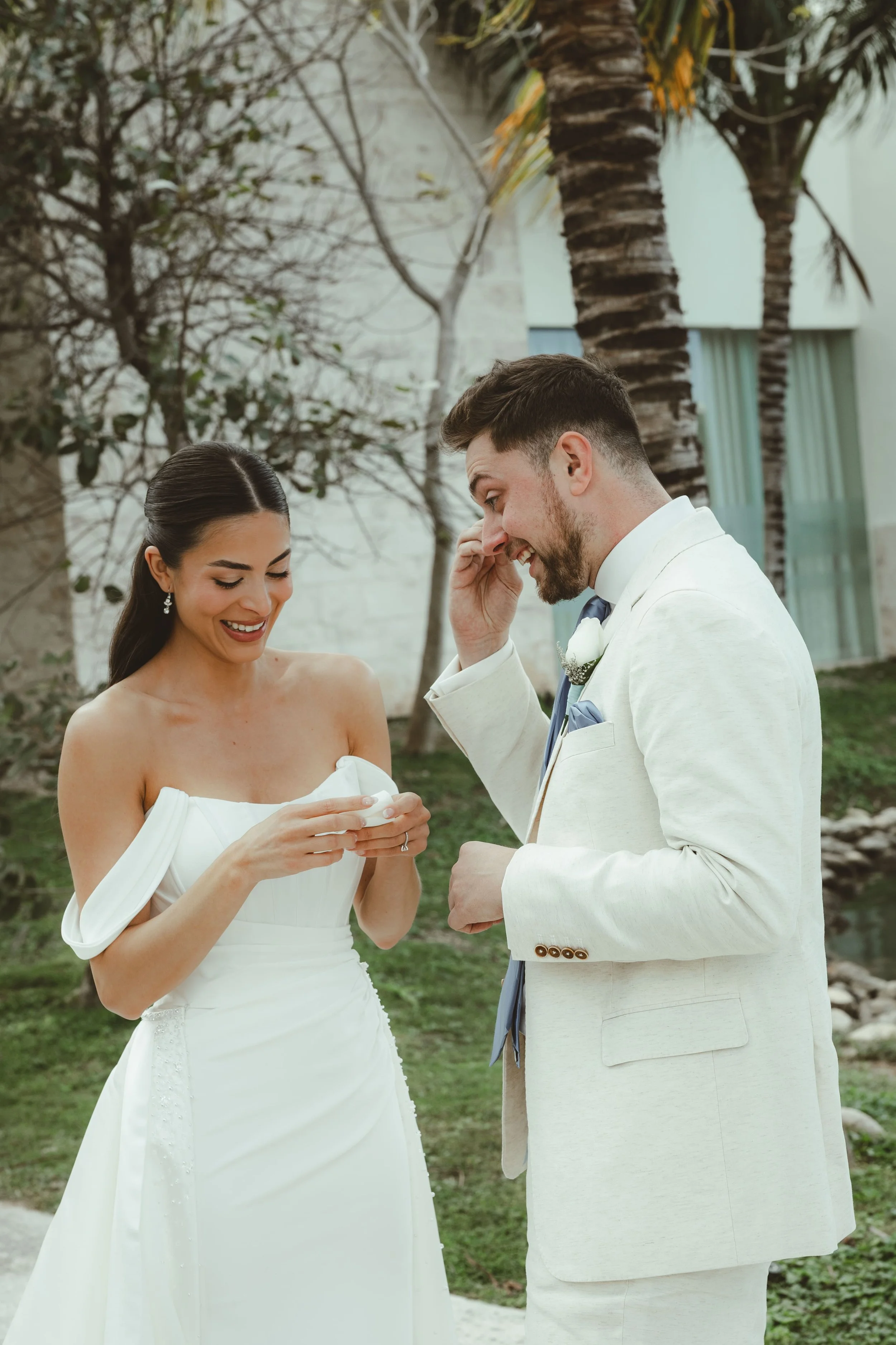A bride and groom exchange rings outdoors, smiling, with palm trees and a building in the background.
