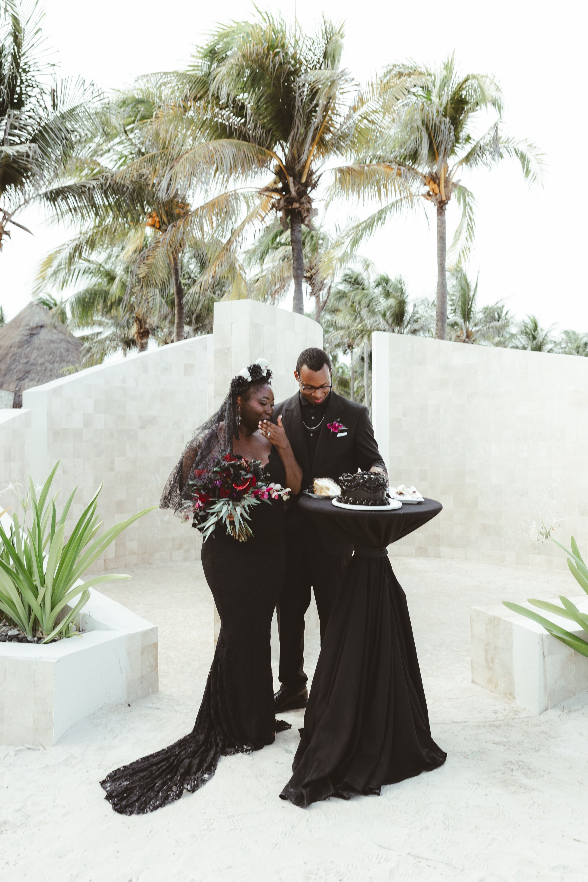 A couple celebrating a wedding outdoors in a tropical setting, with palm trees in the background. They are dressed in formal attire, with the woman in a black lace dress and veil, holding a bouquet, and the man in a black suit with a boutonnière. The