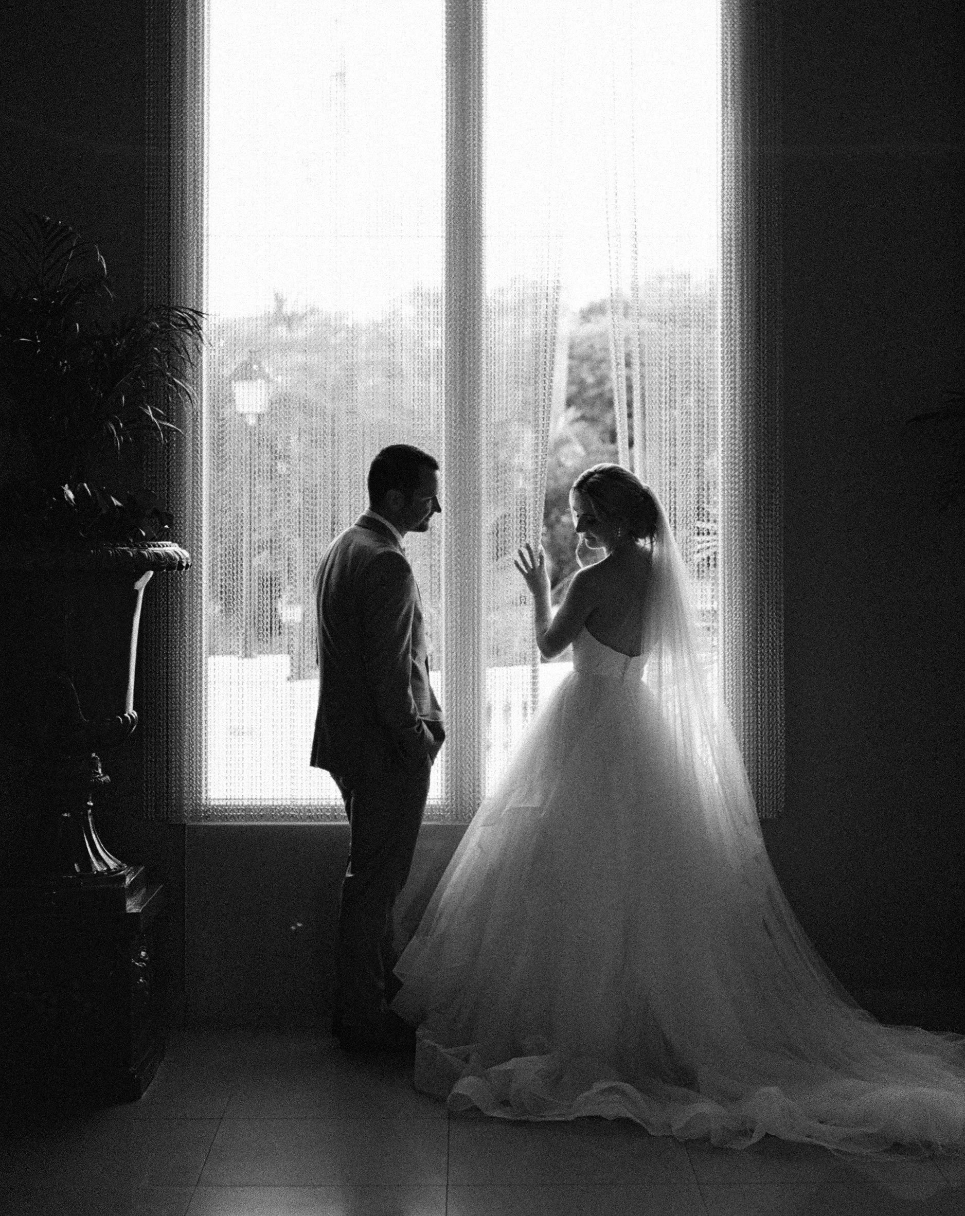 Black and white photo of a bride and groom standing in front of window with curtains, possibly during their wedding or a portrait session.