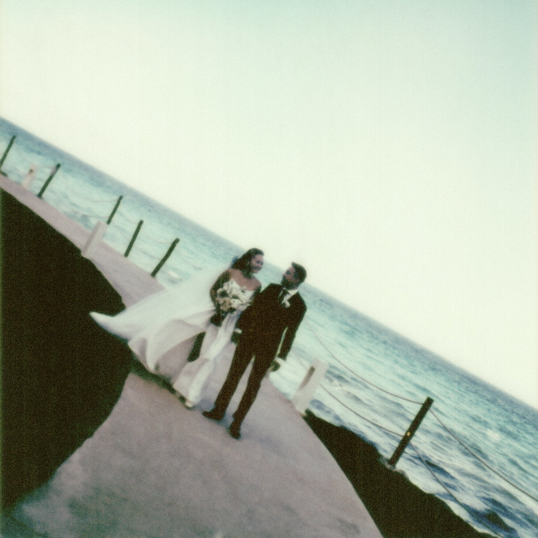 Bride and groom in formal attire, a man and a woman, standing on a beach with a pier and ocean in the background, at a wedding in Mexico.
