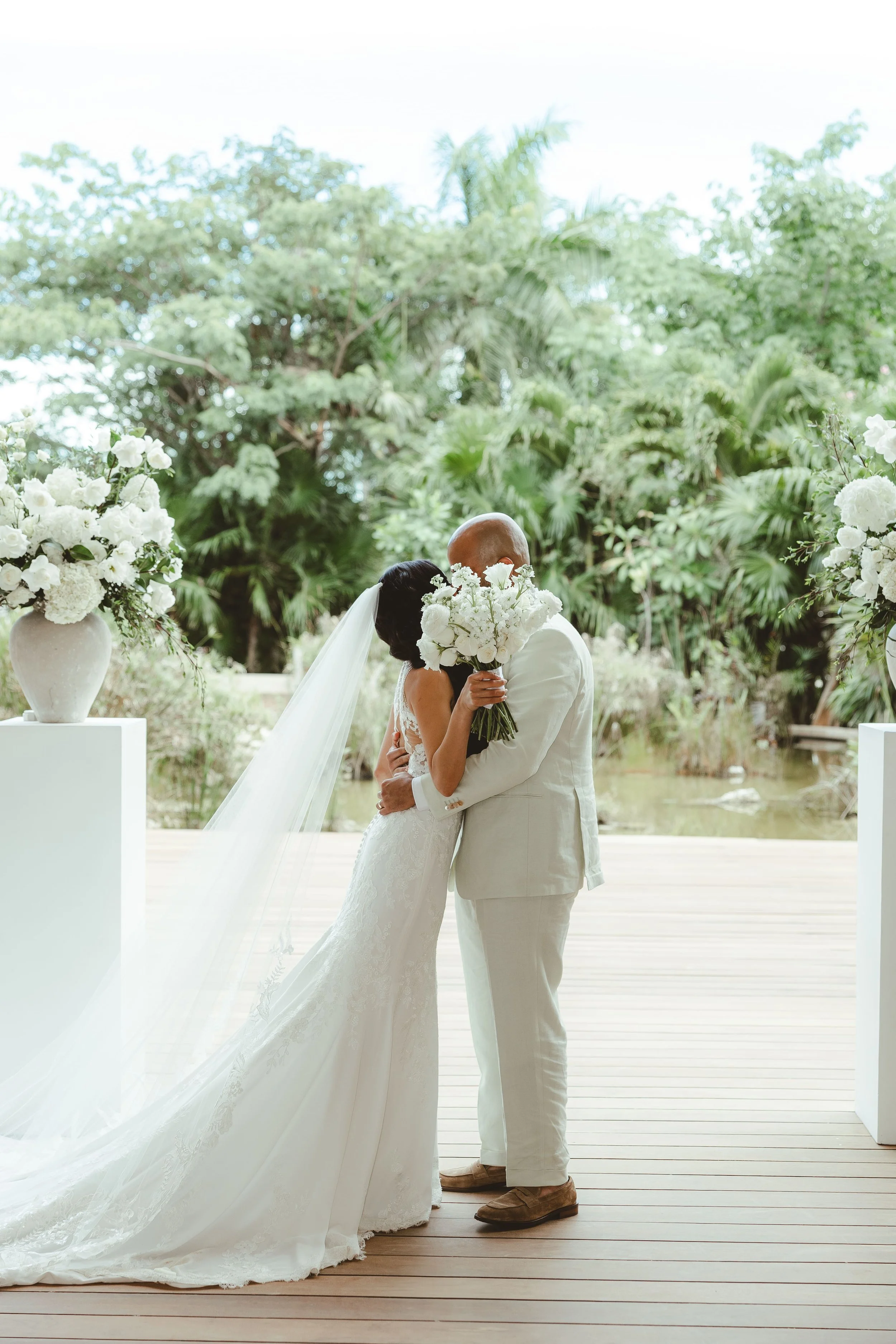 A bride and groom share a kiss during their wedding ceremony outdoors, surrounded by lush green trees and decorated with white flowers in white vases.