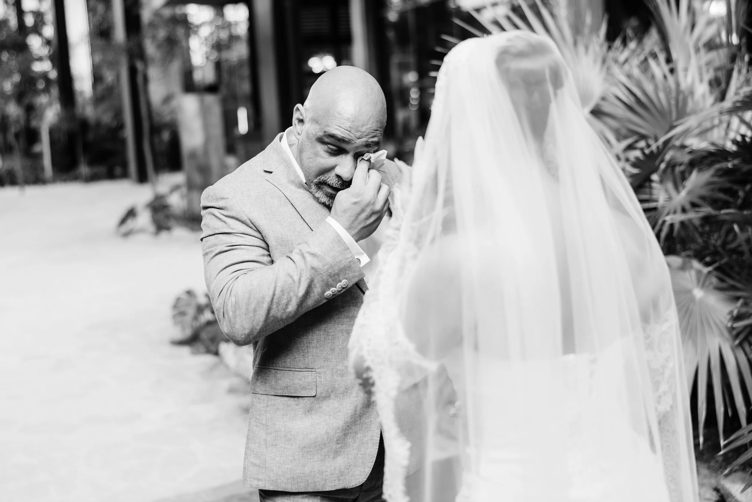 A man in a suit, appearing emotional, wiping tears with a tissue, standing near a bride dressed in a white gown with a veil, at a wedding or similar ceremony in an outdoor setting with trees and plants.