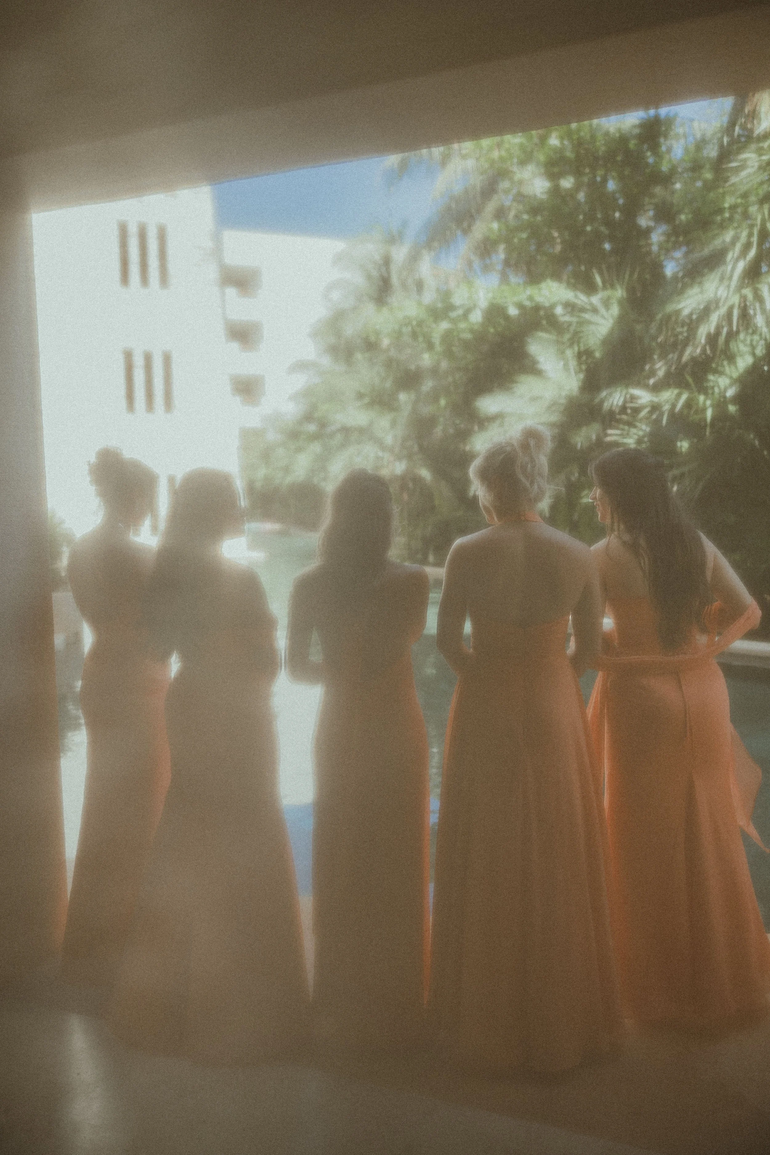 Five women in long dresses standing by a pool, with greenery and a white building and bright sky in the background.