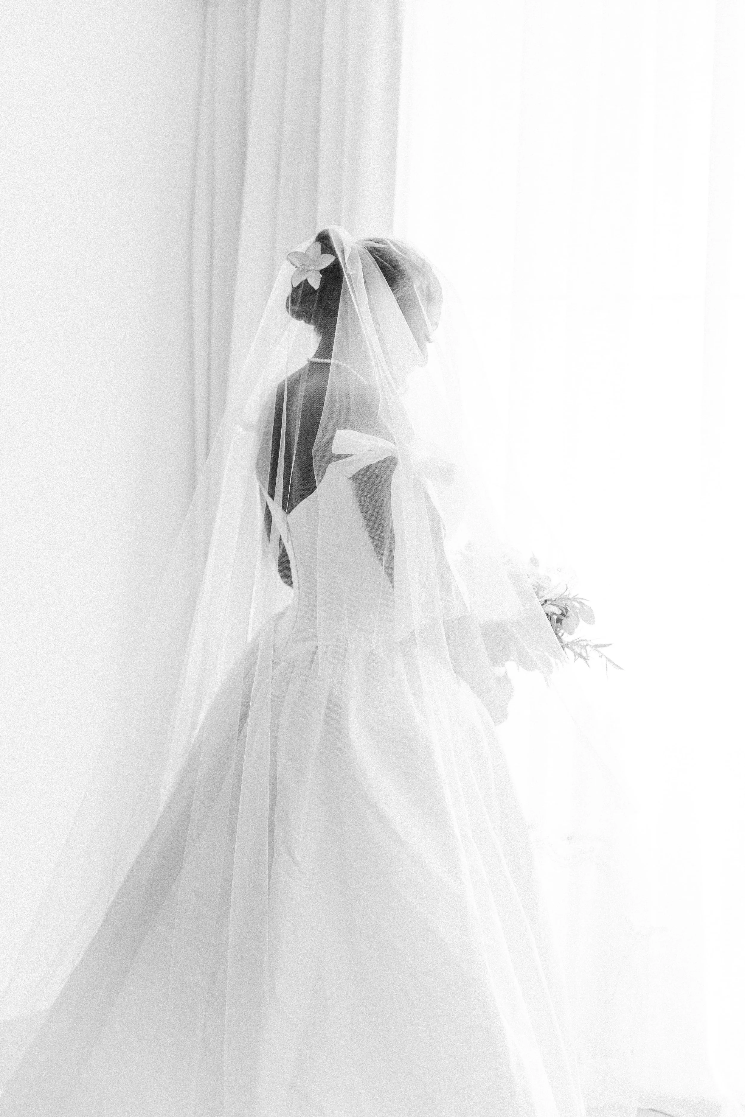 A woman wearing a wedding dress and a veil, standing in front of a light-colored curtain.