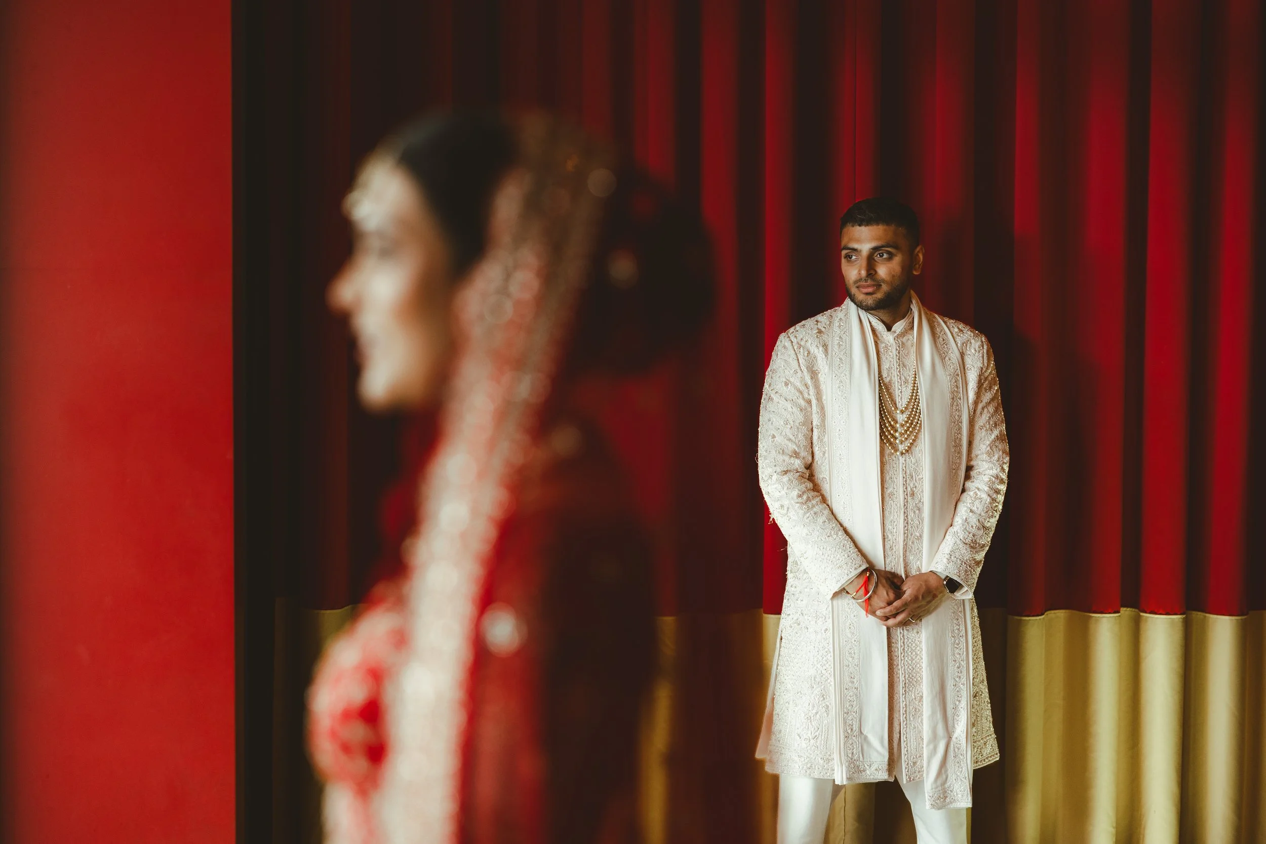 A man dressed in traditional Indian wedding attire stands in front of a red curtain, his hands clasped in front of him, gazing to his right with a calm expression. A woman in ornate attire and jewelry is blurred in the foreground, facing him.
