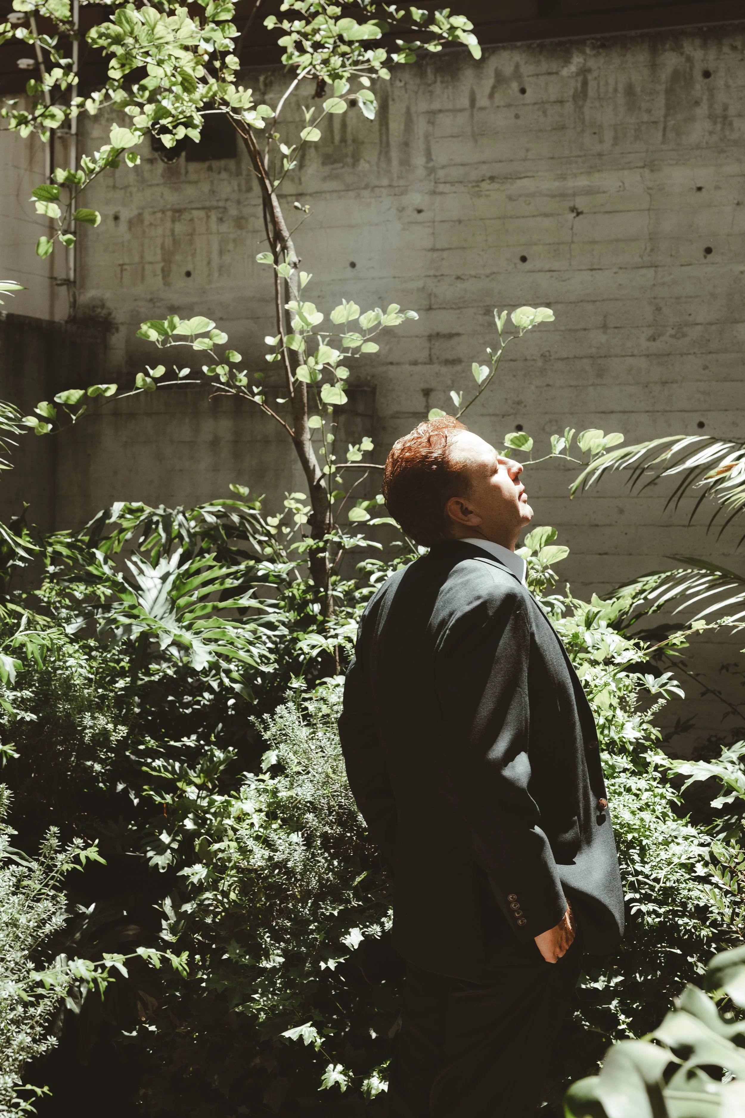 A man with reddish hair, wearing a black suit, is standing outdoors surrounded by lush green plants. He is looking up towards the sky with his eyes closed, appearing relaxed and contemplative, with sunlight illuminating his face.