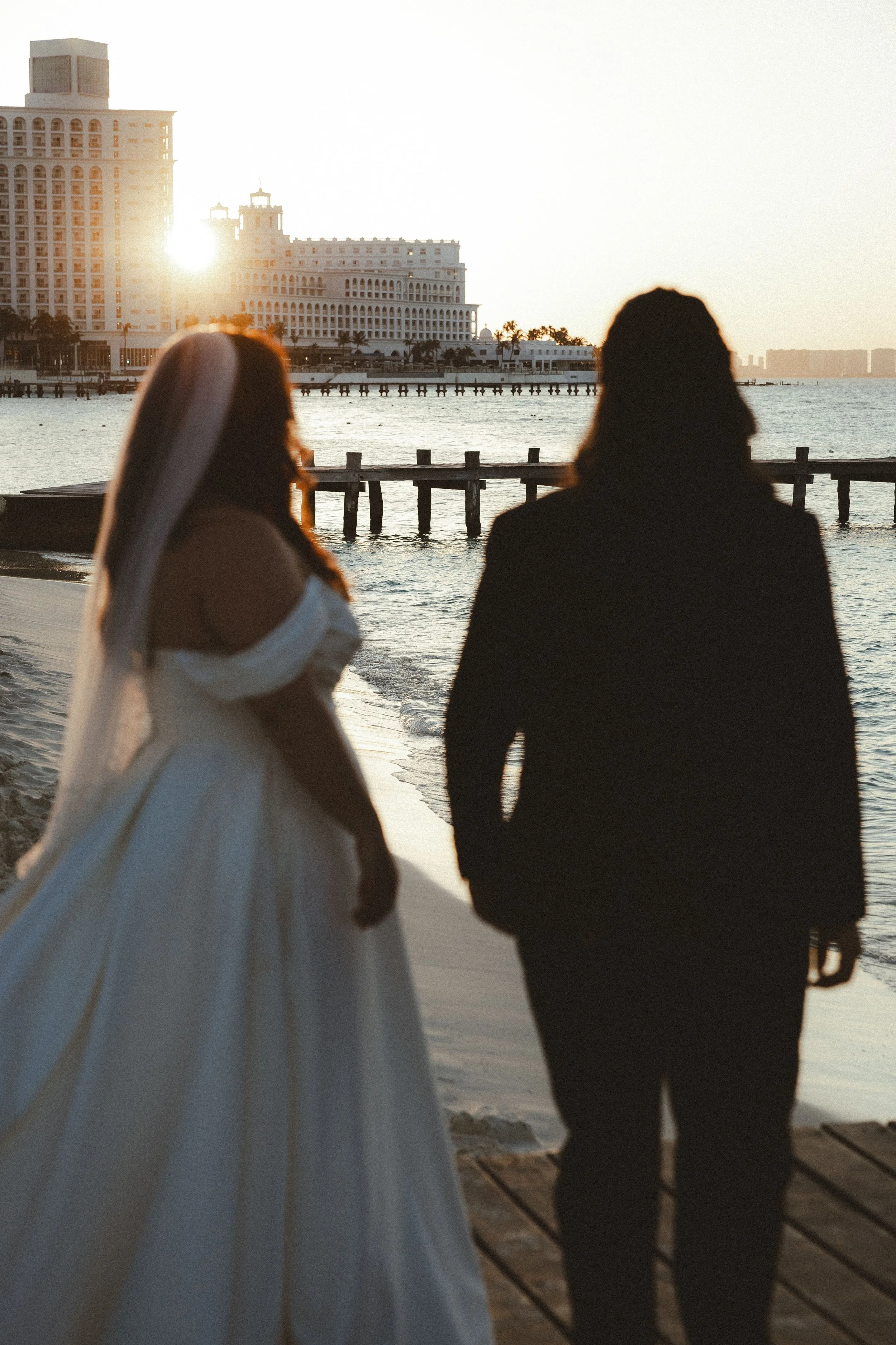 Two women walking on a beach at sunset, with a harbor, a pier, and a large white hotel in the background.