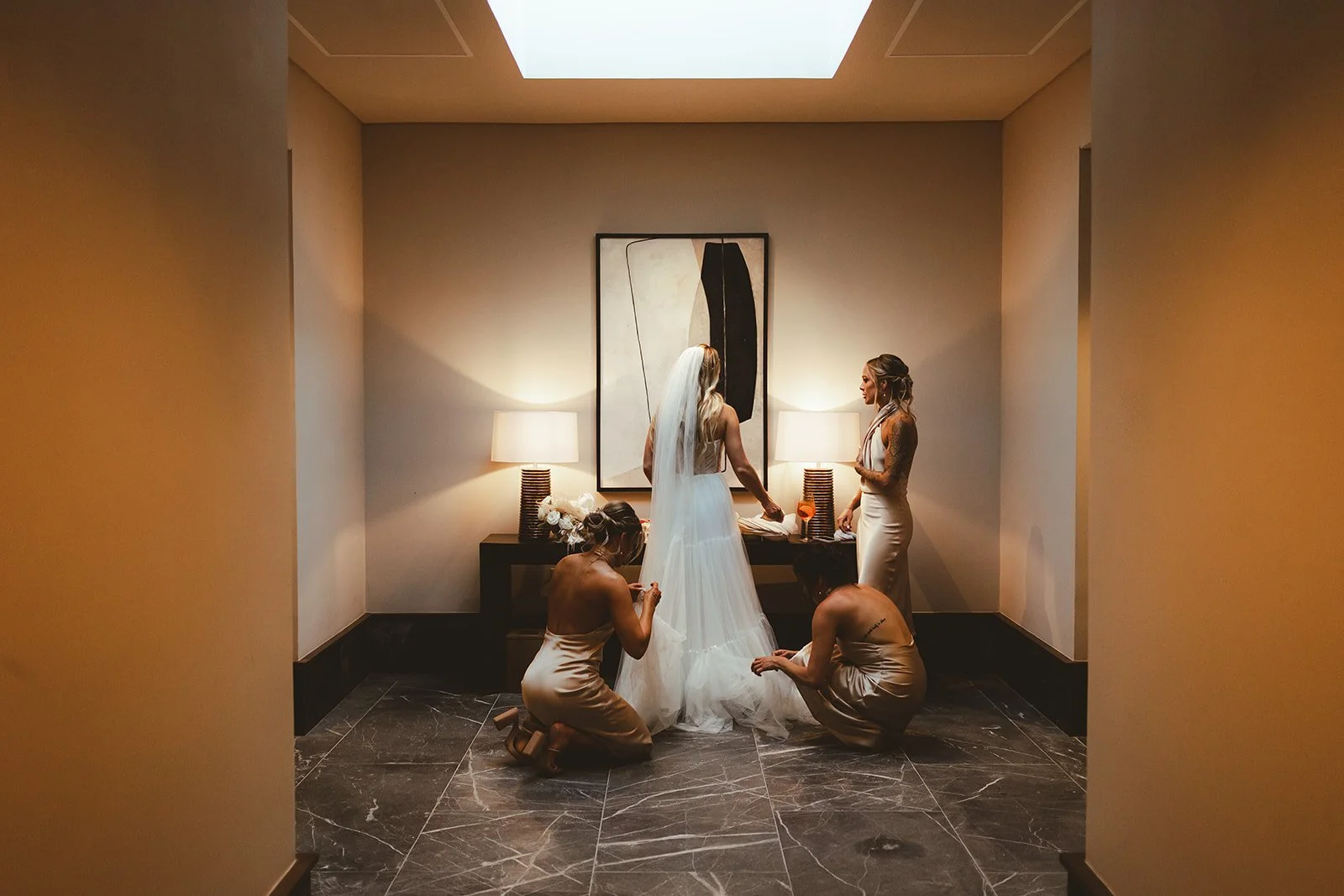 Bride in a wedding dress with veil being helped by two bridesmaids in a hotel room, with a painting and two lamps on a table behind them.