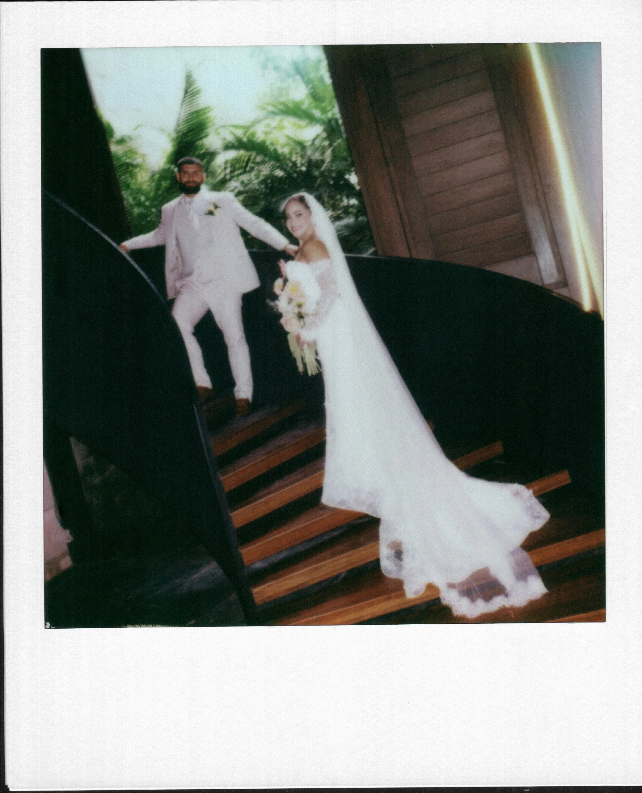 A wedding photo of a couple standing on a staircase outdoors, with lush green trees in the background. The man is dressed in a light-colored suit and the woman in a white wedding gown holding a bouquet at Secrets Moxche wedding in Playa del Carmen.
