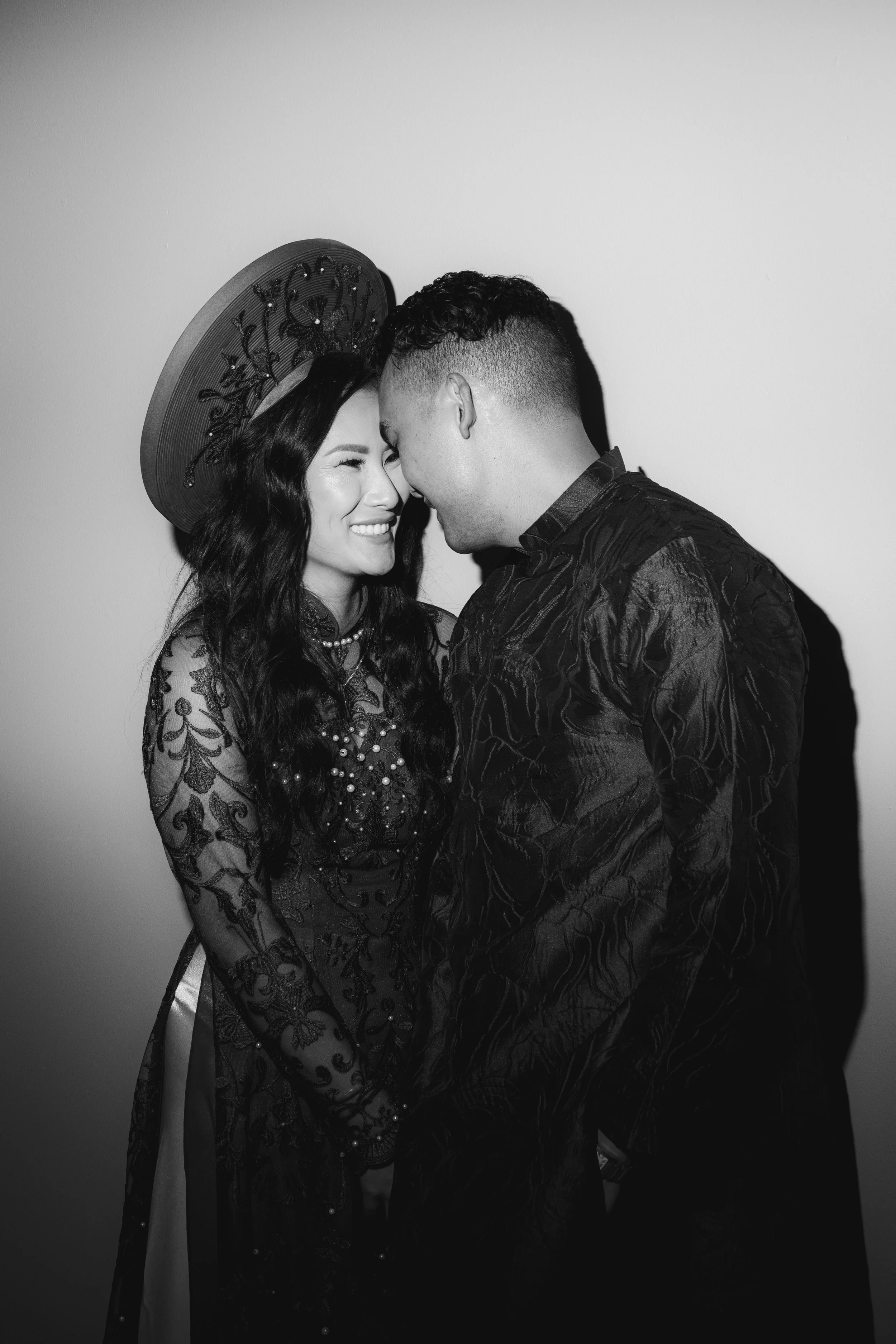 Black and white photo of a smiling woman with long dark hair wearing a large hat with floral embroidery and a lace dress, and a man with short curly hair and a patterned jacket, leaning their foreheads together in an intimate pose.