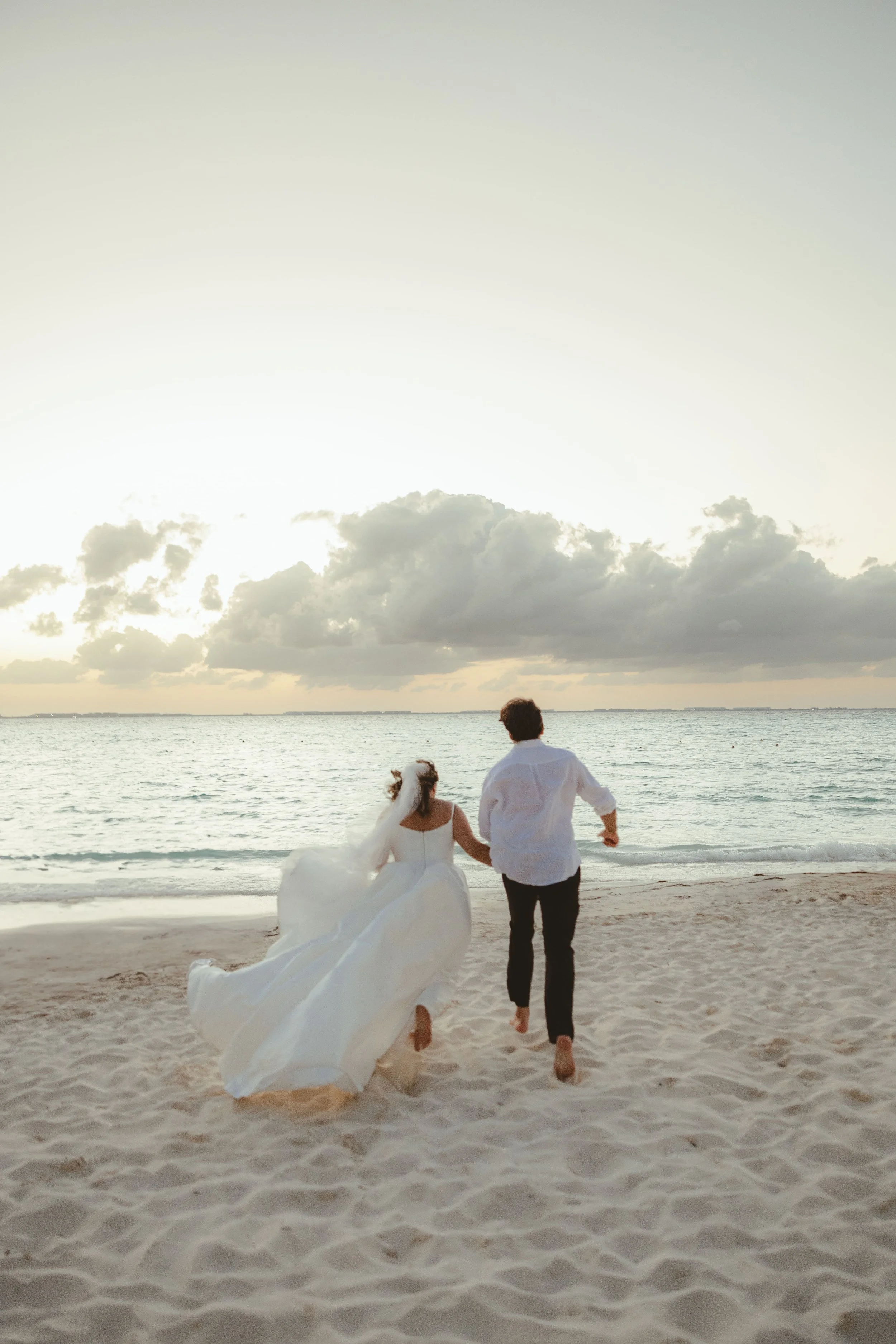 A bride and groom walking hand in hand on the beach during sunset, with the bride in a white wedding dress and veil, and the groom in black pants and a white shirt.