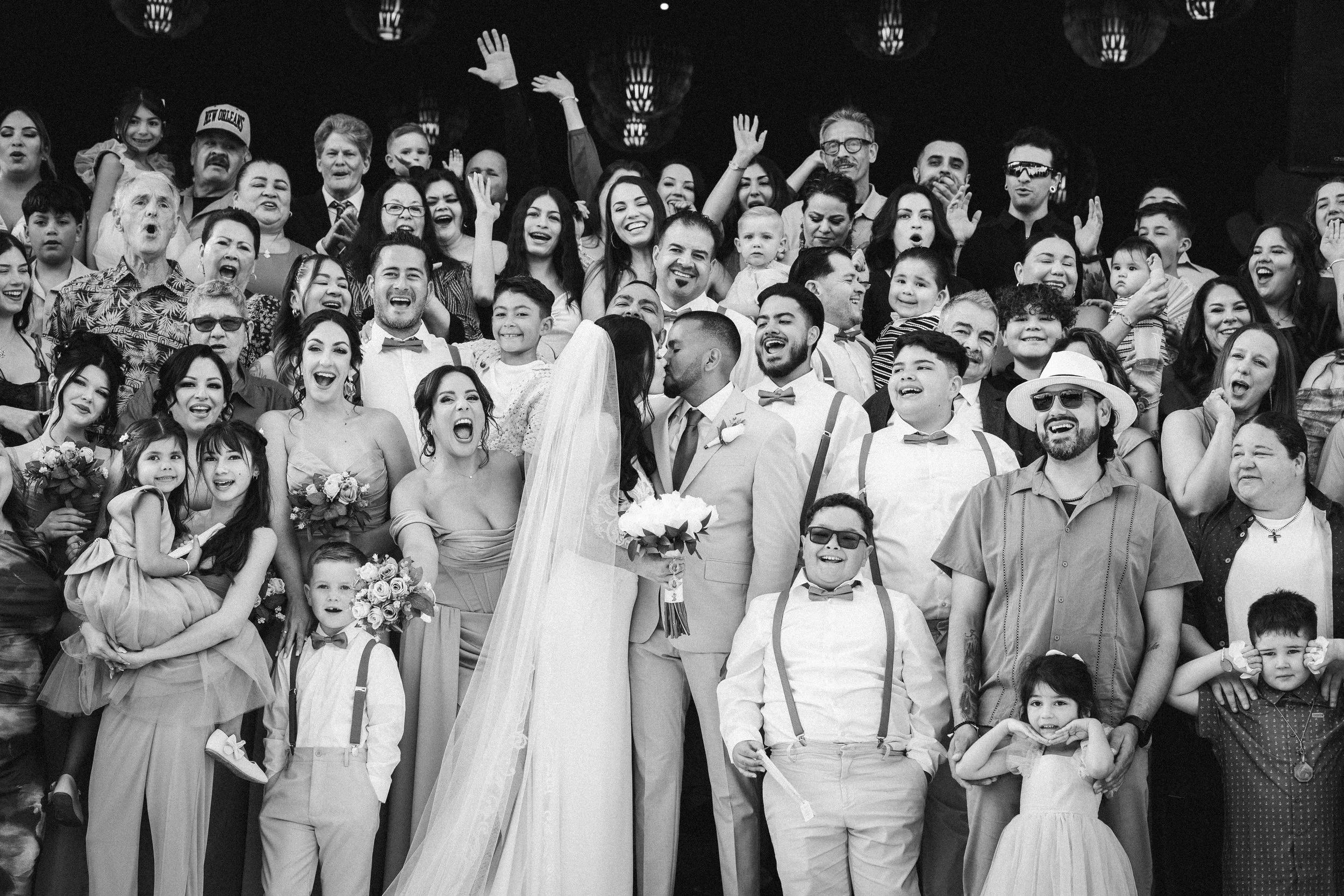 A large group of people at a wedding, smiling and celebrating, with some raising their hands, wearing formal and casual attire, and holding flowers.