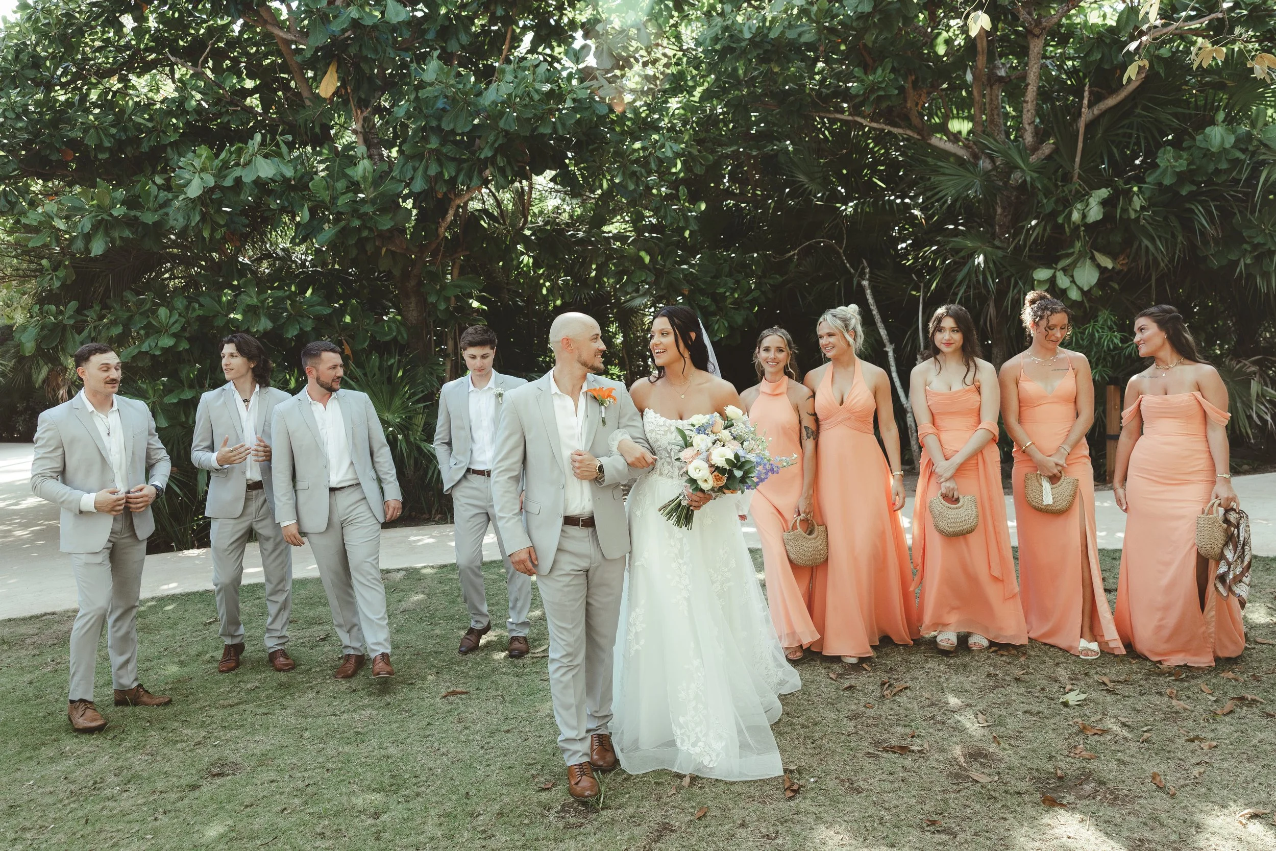 A wedding party walking outdoors on grass, with large green trees in the background. The bride in a white gown with dark hair, the groom in a light gray suit with a boutonniere, and six bridesmaids in peach dresses follow behind them, all smiling.