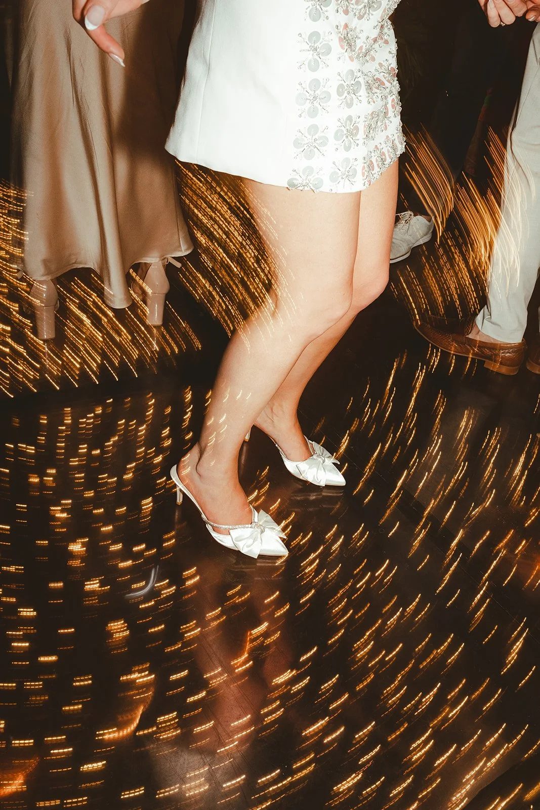 A woman wearing white high heels with bows and a white floral dress is standing on a shiny black floor with swirling lights around.