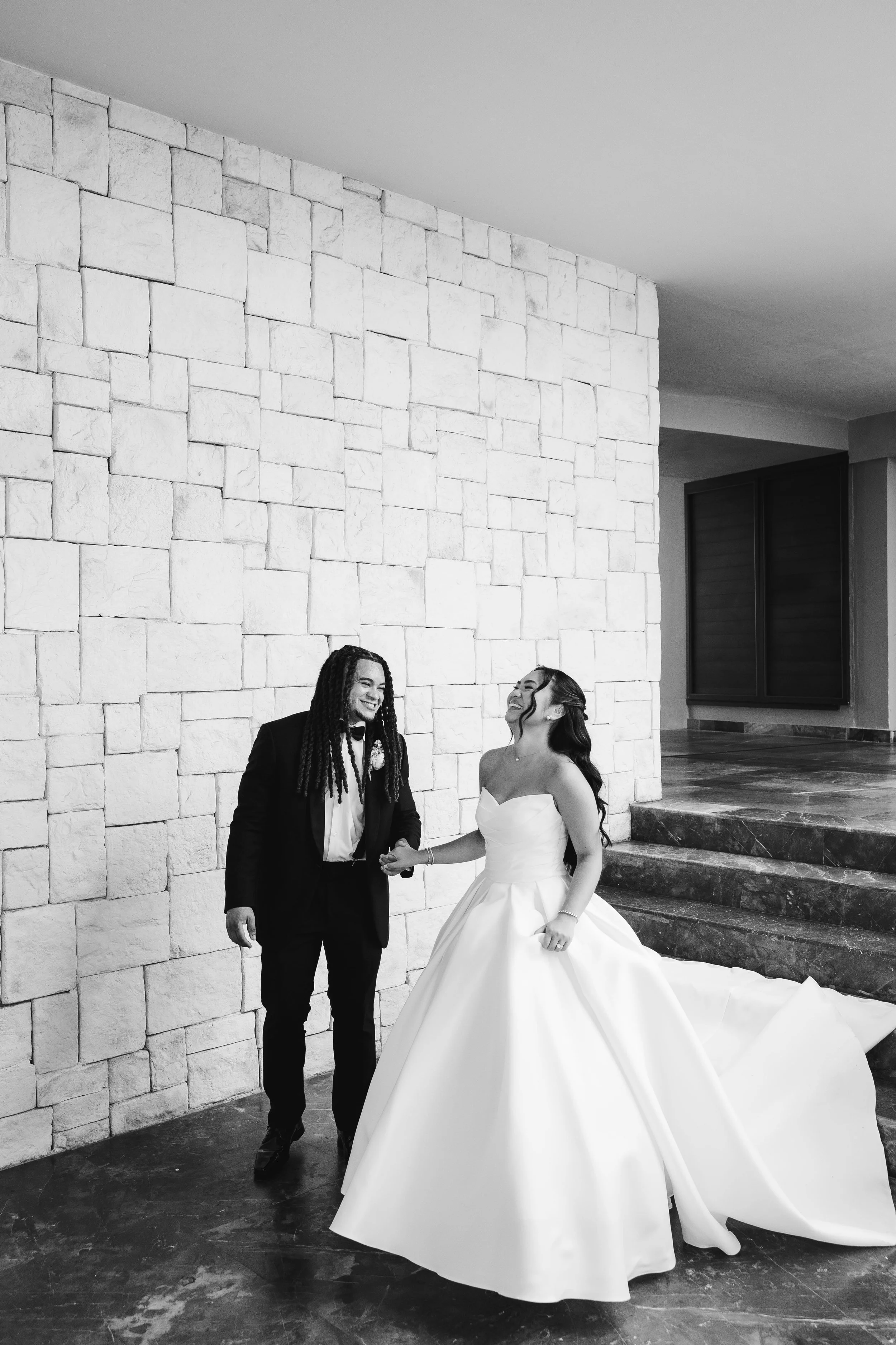 A black-and-white photo of a couple at their wedding, holding hands and smiling at each other indoors against a stone wall and window. The bride is wearing a strapless ball gown, and the groom is in a formal suit.