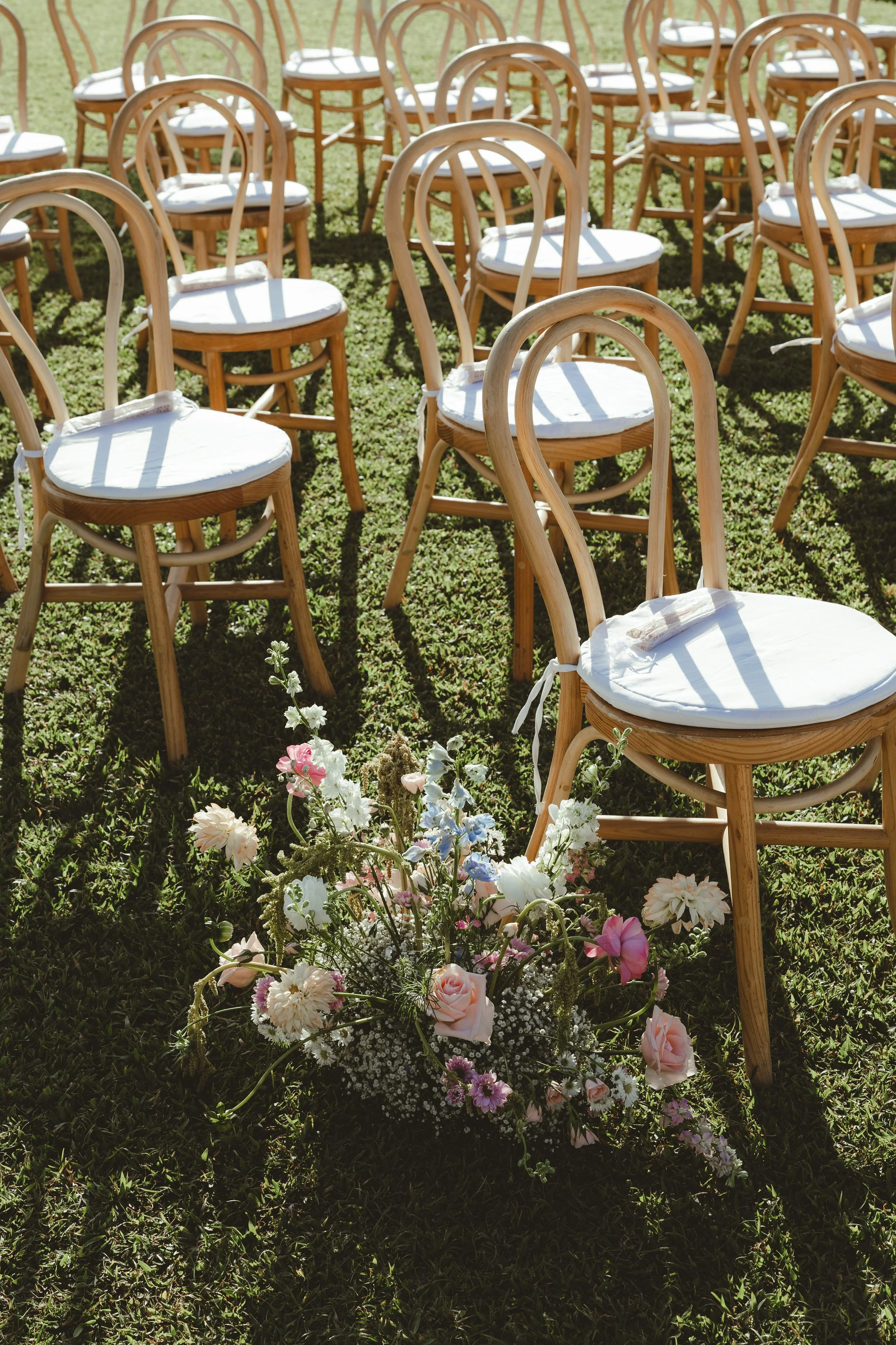 Empty wooden chairs with white cushions are arranged outdoors on a grassy lawn, with a colorful flower bouquet in the foreground.