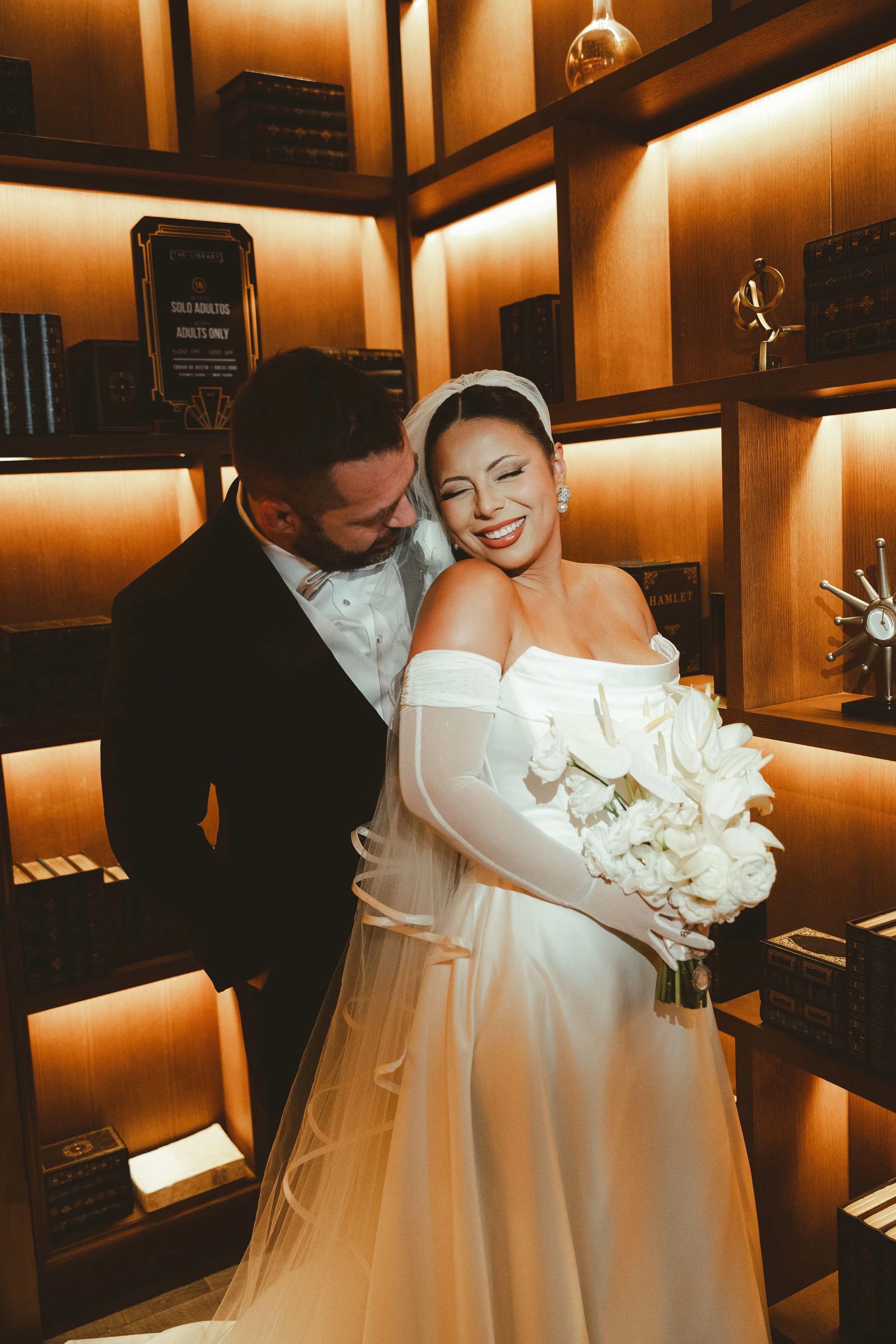A bride smiling and holding a bouquet of white flowers, standing in front of a wooden bookshelf with books and decorative objects, with a groom in a tuxedo close behind her.