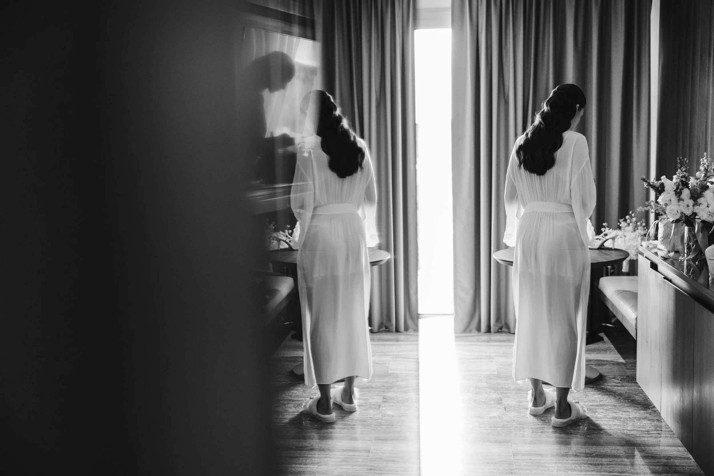 A black and white photo of a woman in a long dress standing in front of a mirror, with her reflection visible. She is in a room with curtains and a bouquet of flowers on a table to her right.