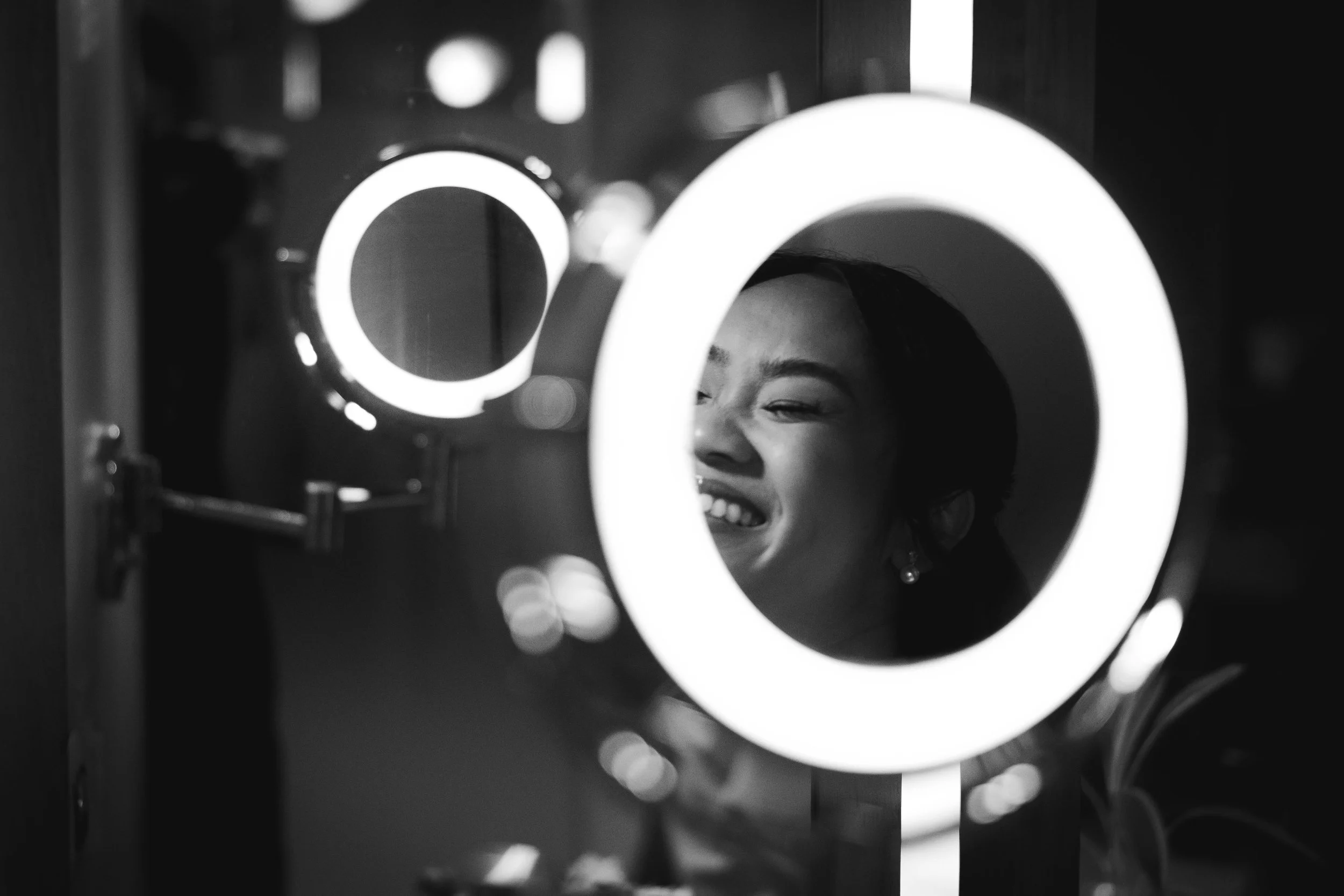 A woman smiling and looking through a circular mirror with LED ring lights, creating a bright, glowing frame around her face.