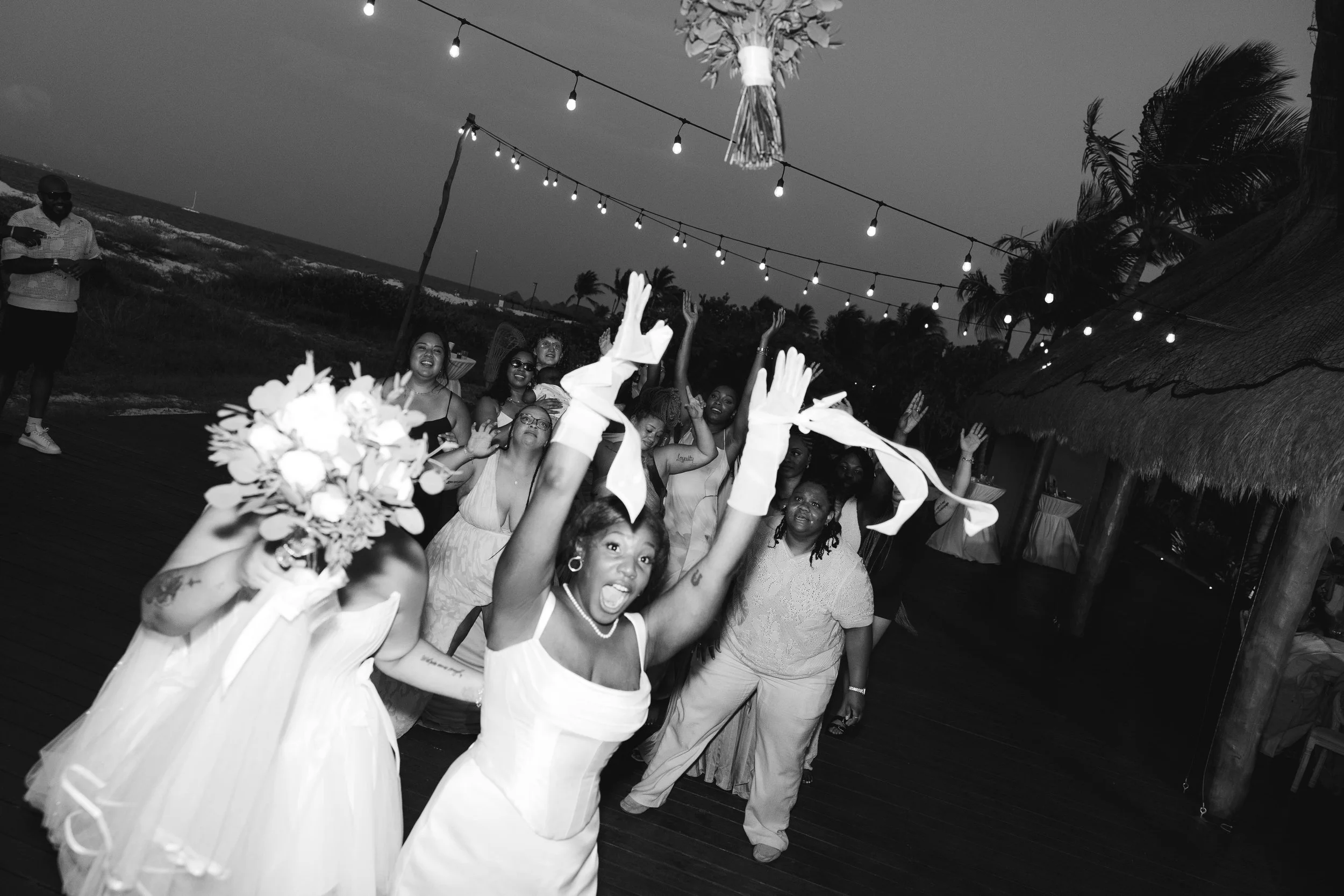 People gathered at an outdoor party during sunset, some dancing and others taking photos, with string lights hanging above and a thatched roof on the right.