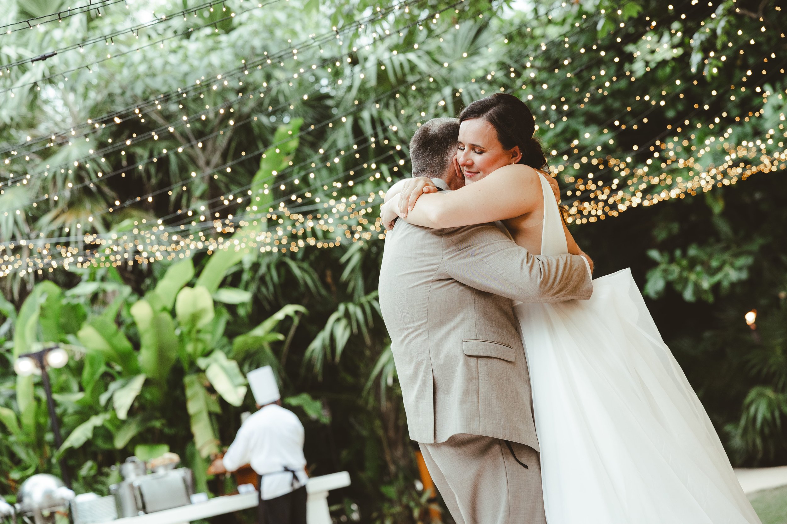 A bride and groom hugging during their wedding reception outdoors, with string lights and lush greenery in the background.