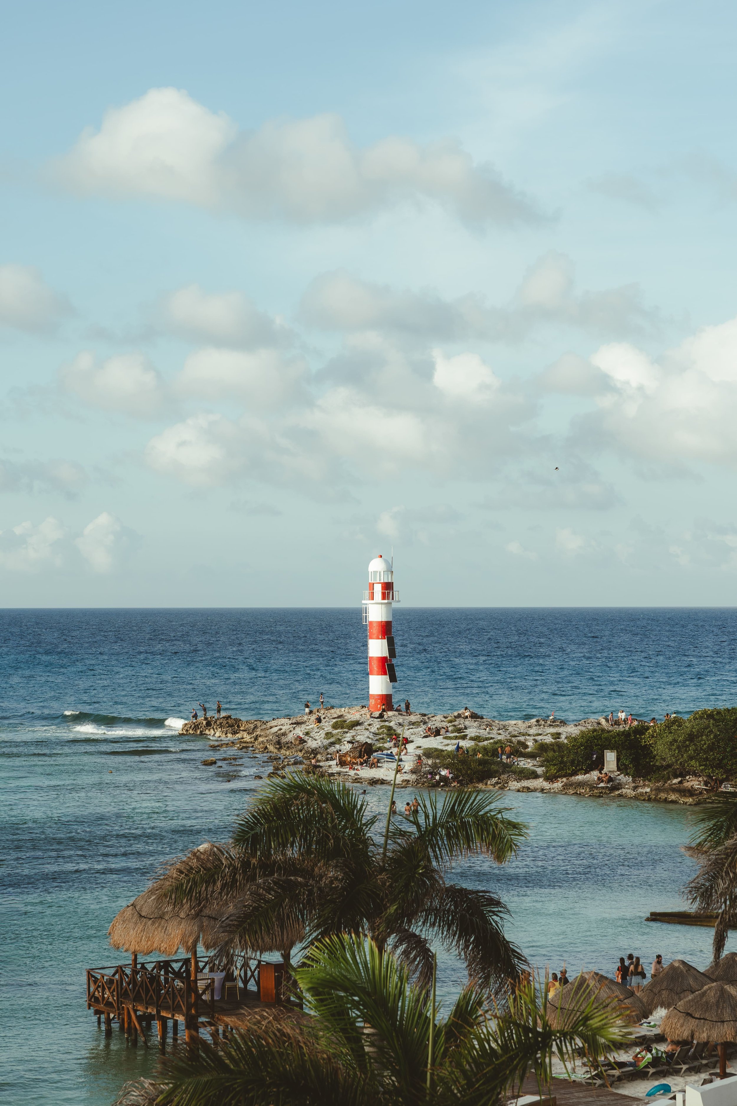 A lighthouse with red and white stripes on a rocky promontory near the ocean, with people relaxing and enjoying the view. Palm trees and thatched-roof structures are in the foreground, under a partly cloudy sky.