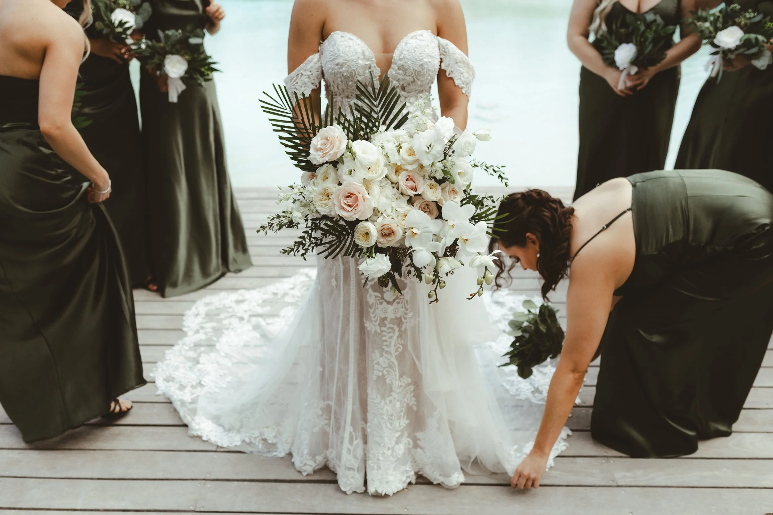 A bride in a lace wedding dress with off-the-shoulder sleeves holds a large bouquet of white and blush flowers with green leaves, surrounded by bridesmaids dressed in dark gowns, on a wooden deck by the water.