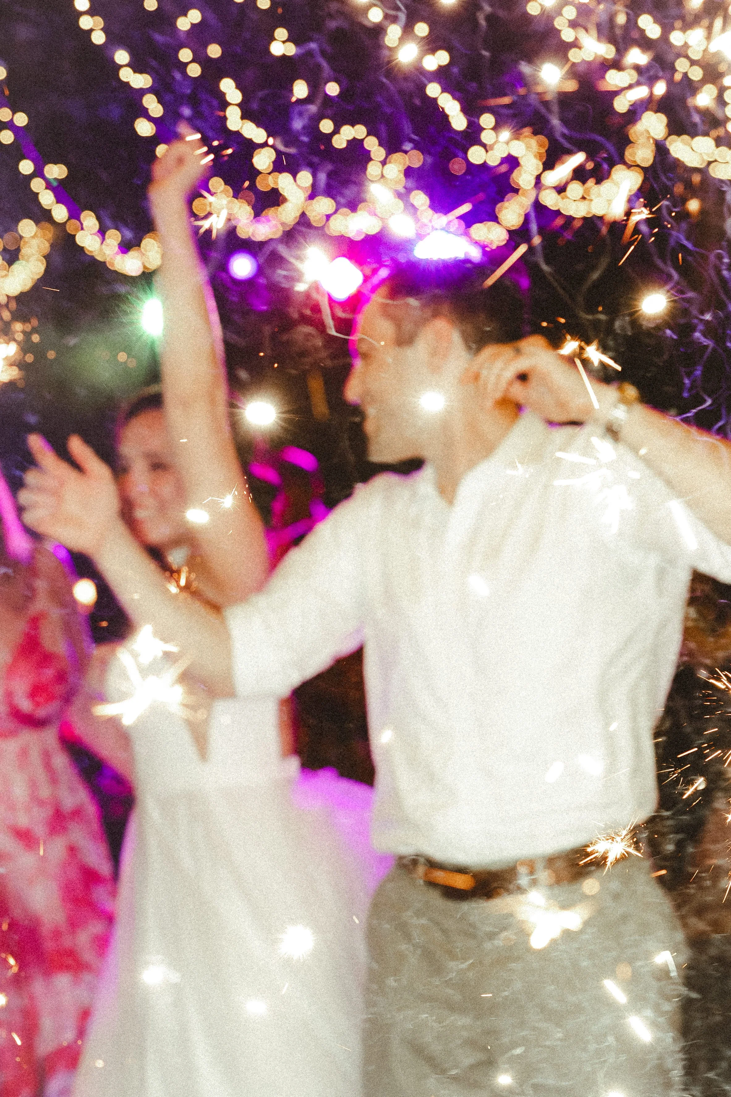 People celebrating with sparklers at a party or event, with colorful lights and bokeh effect in the background.