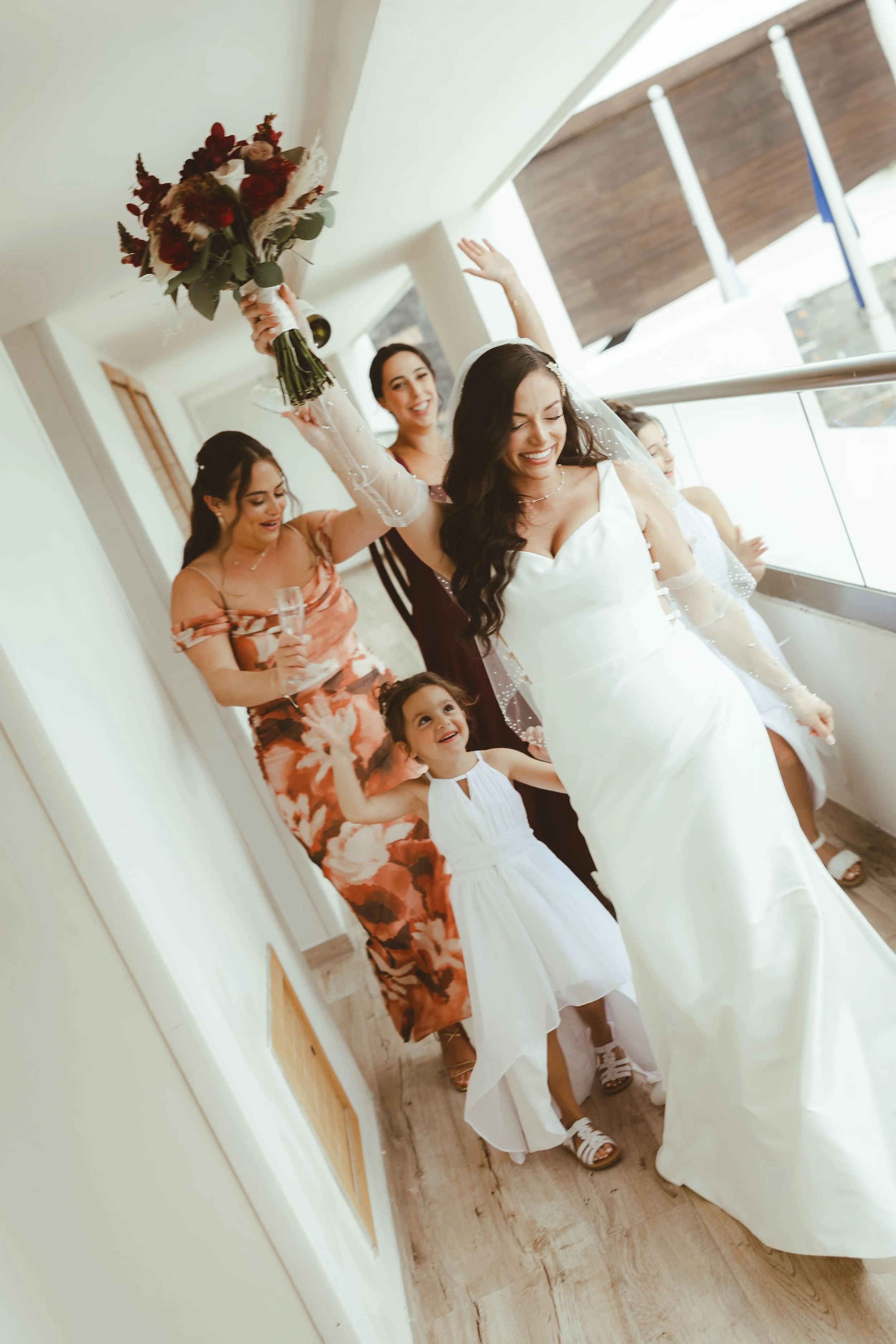 A bride in a white wedding dress joyfully waves while holding a bouquet of red and white flowers above her head. She is surrounded by women and a young girl, all smiling and celebrating, in a well-lit hallway with large windows and wooden flooring.