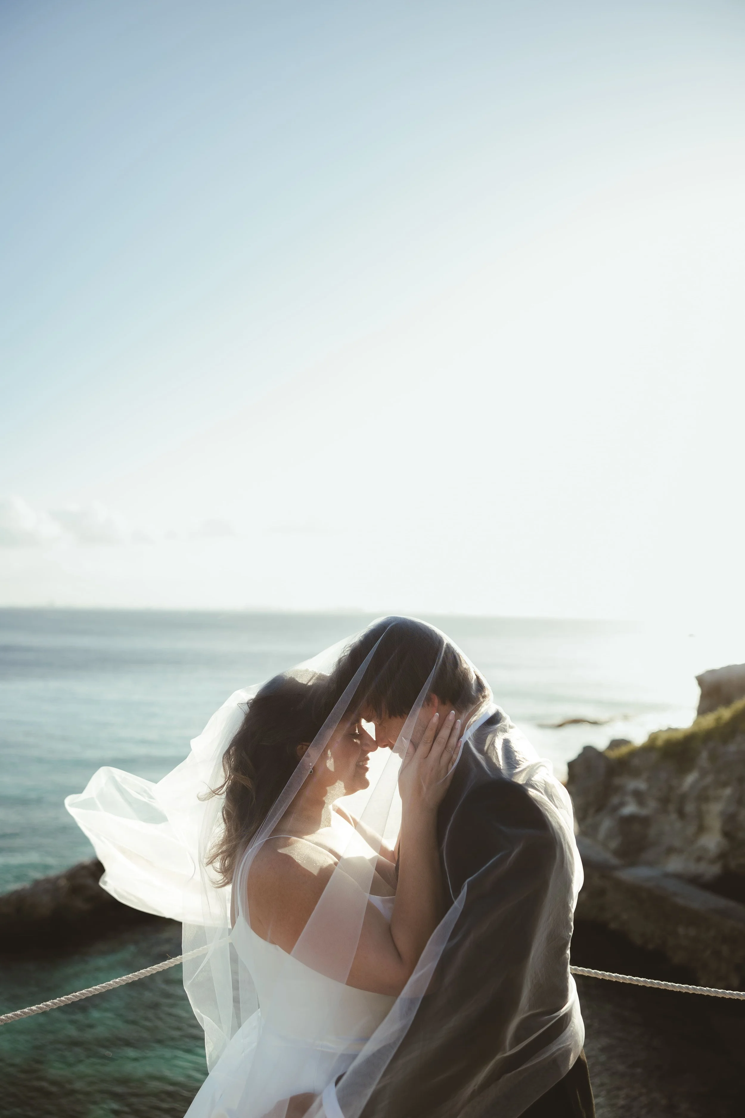 A bride and groom sharing a kiss on the beach, with the ocean and sky in the background, during sunset.