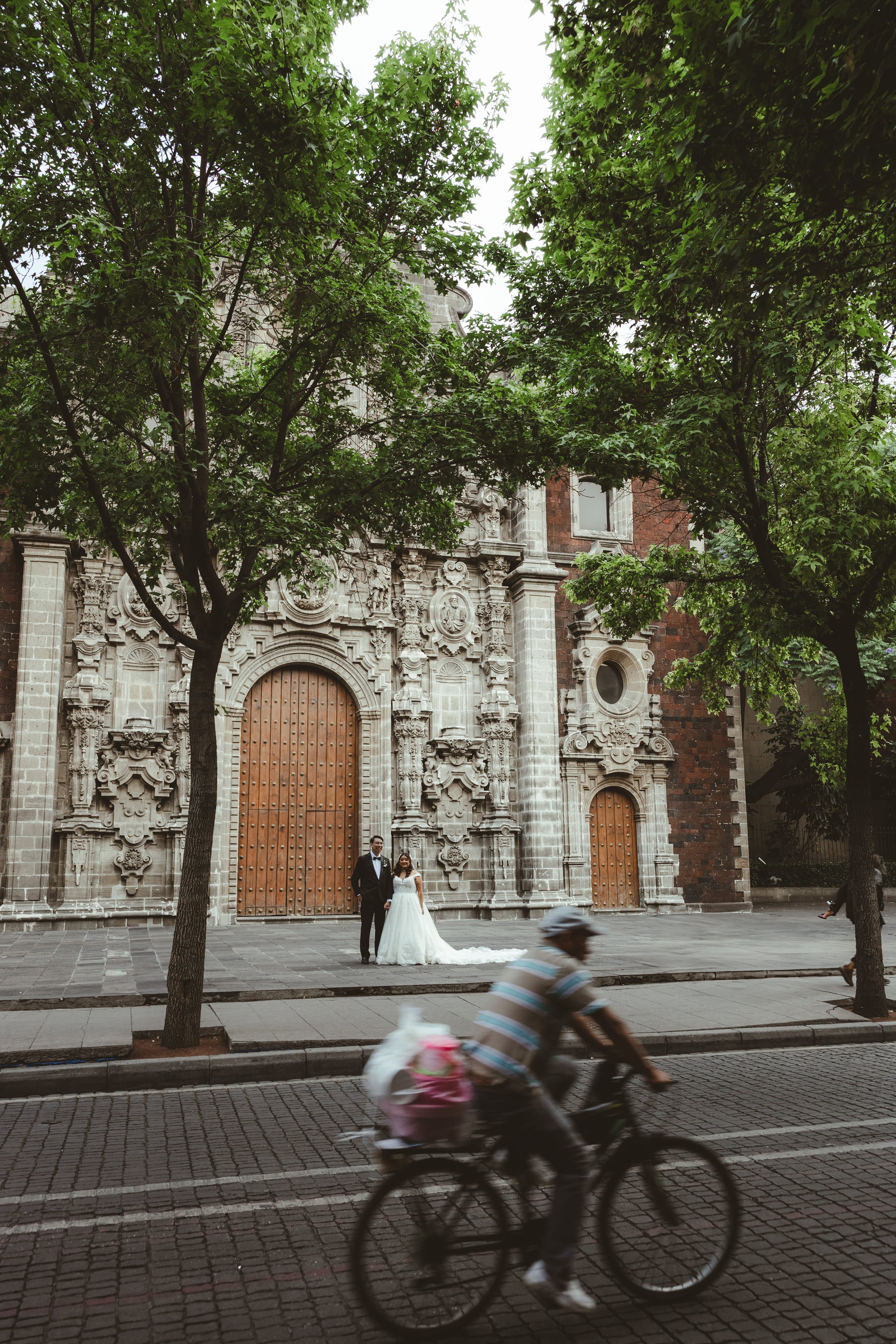A bride and groom standing in front of a historic building with ornate stone details and large wooden doors, while a cyclist passes by in the foreground on a city street lined with trees.