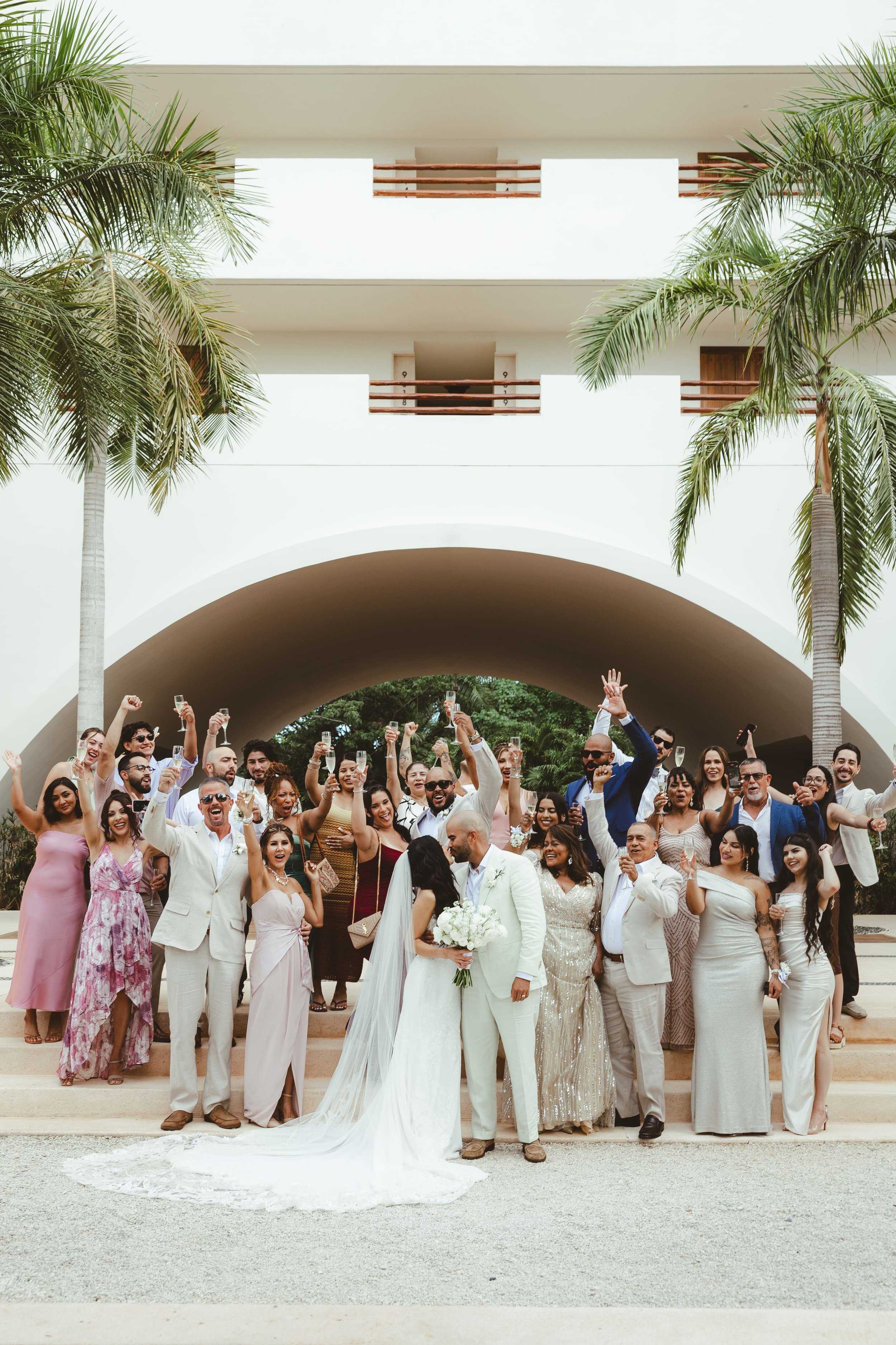 A large group of people dressed in formal attire gathered outdoors, with many raising champagne glasses and smiling. Two women in wedding dresses are in the center, indicating it is a wedding celebration. The backdrop features a modern archway.