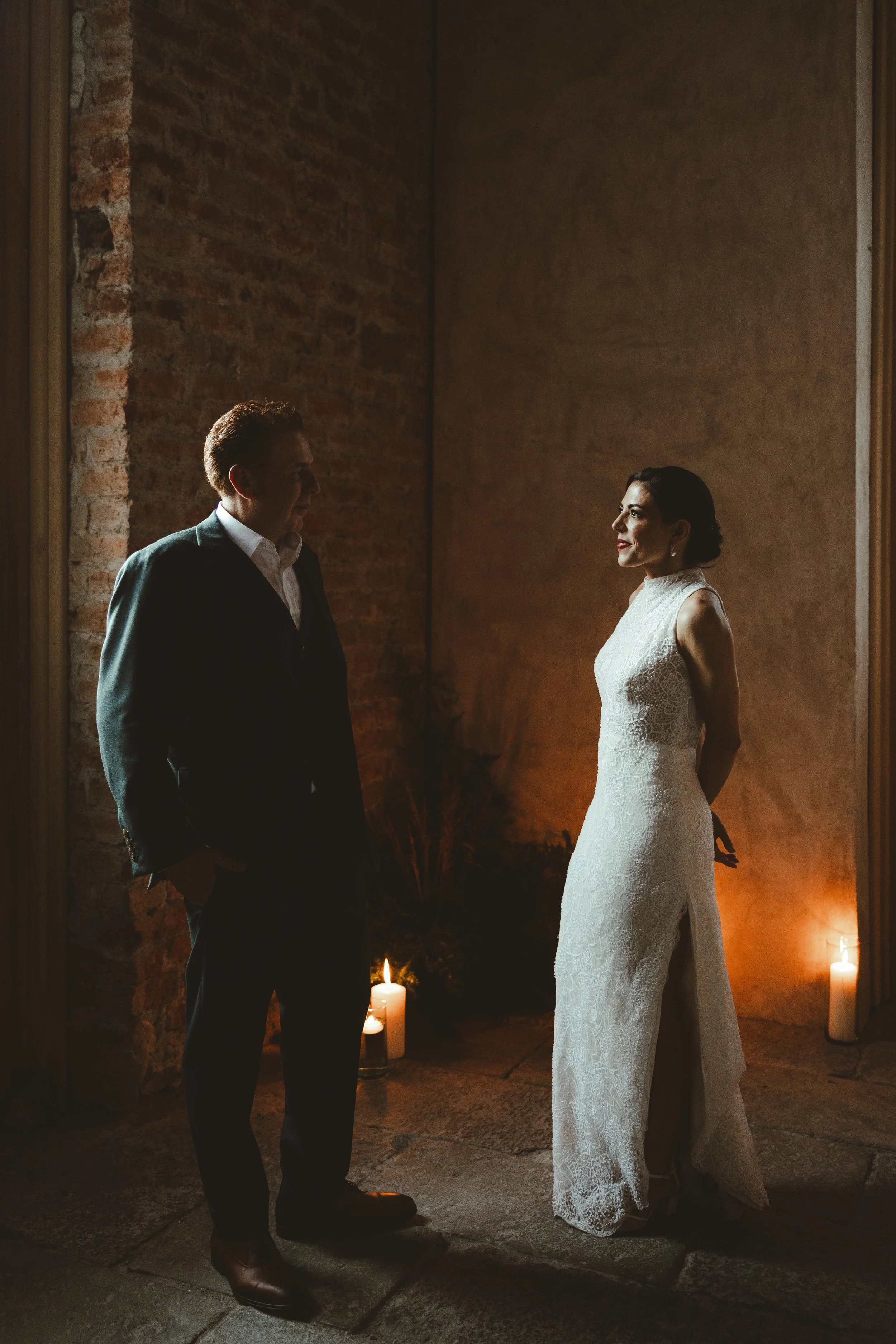 A couple in formal attire standing indoors at night, facing each other and smiling. The man is wearing a dark suit and the woman a white lace dress. Candlelight lights the scene with a warm glow.