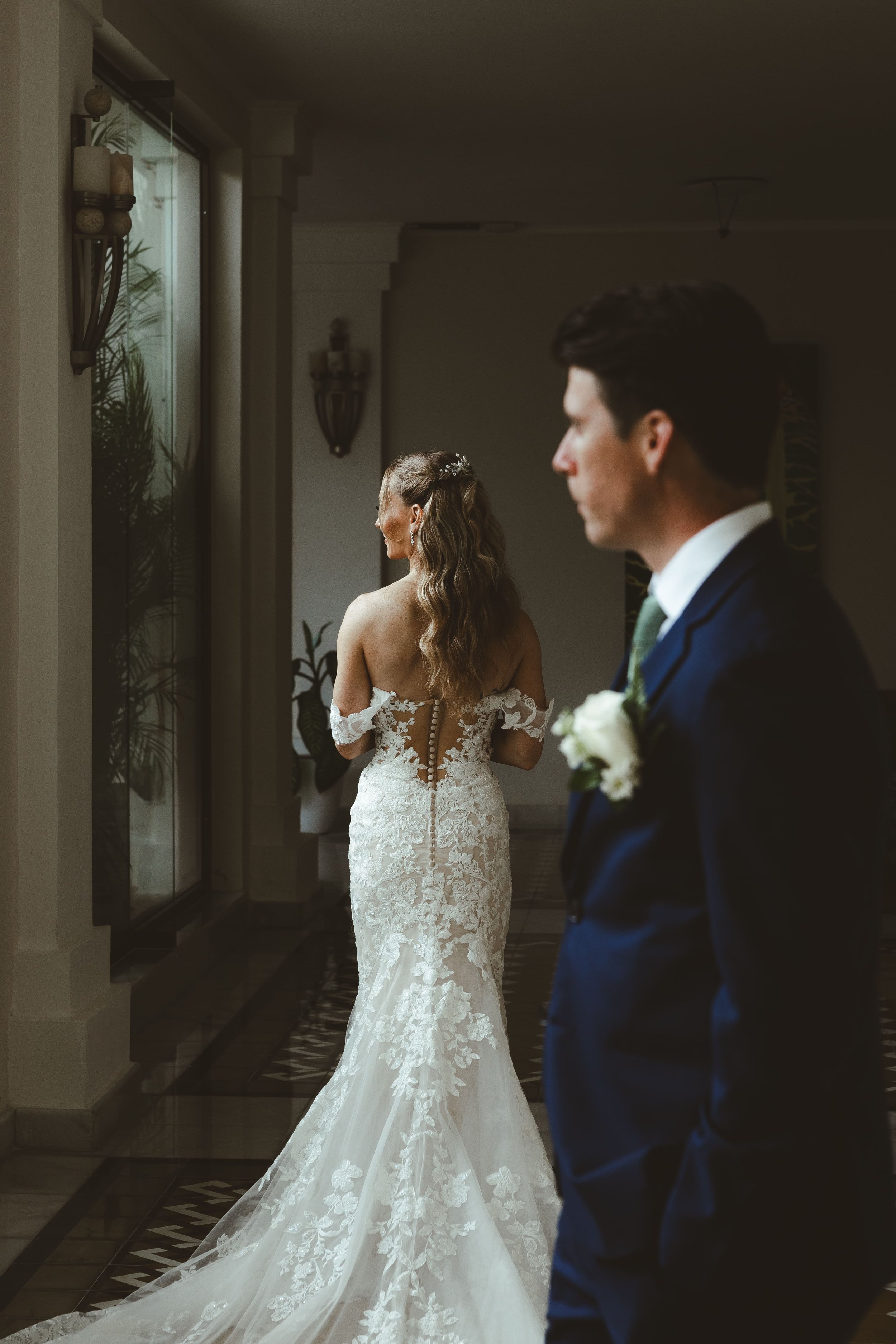 A bride in a white lace wedding dress with a train, standing near a wall and looking out a window, with a groom in a dark suit and white shirt standing nearby.