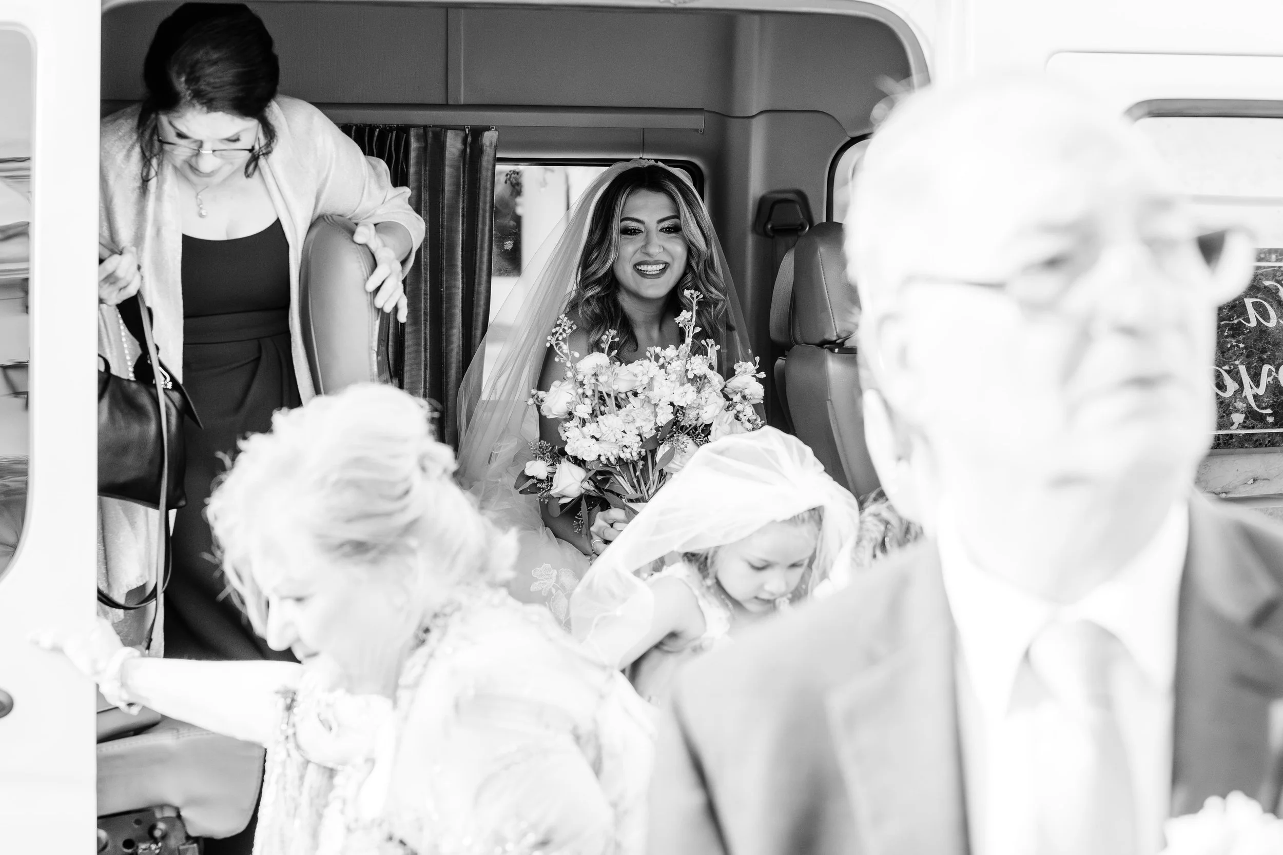 A bride with long hair and a bouquet of flowers sitting inside a vehicle, smiling. Two children in white dresses are seated in front of her, with a woman leaning over from the left side of the vehicle.