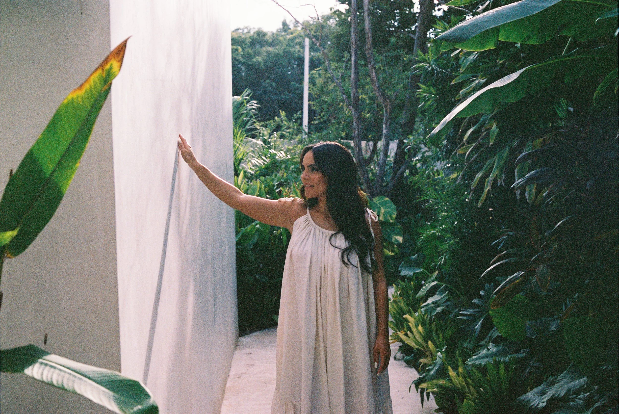 A woman in a light-colored dress smiling and touching a white wall, surrounded by lush green plants in an outdoor setting.