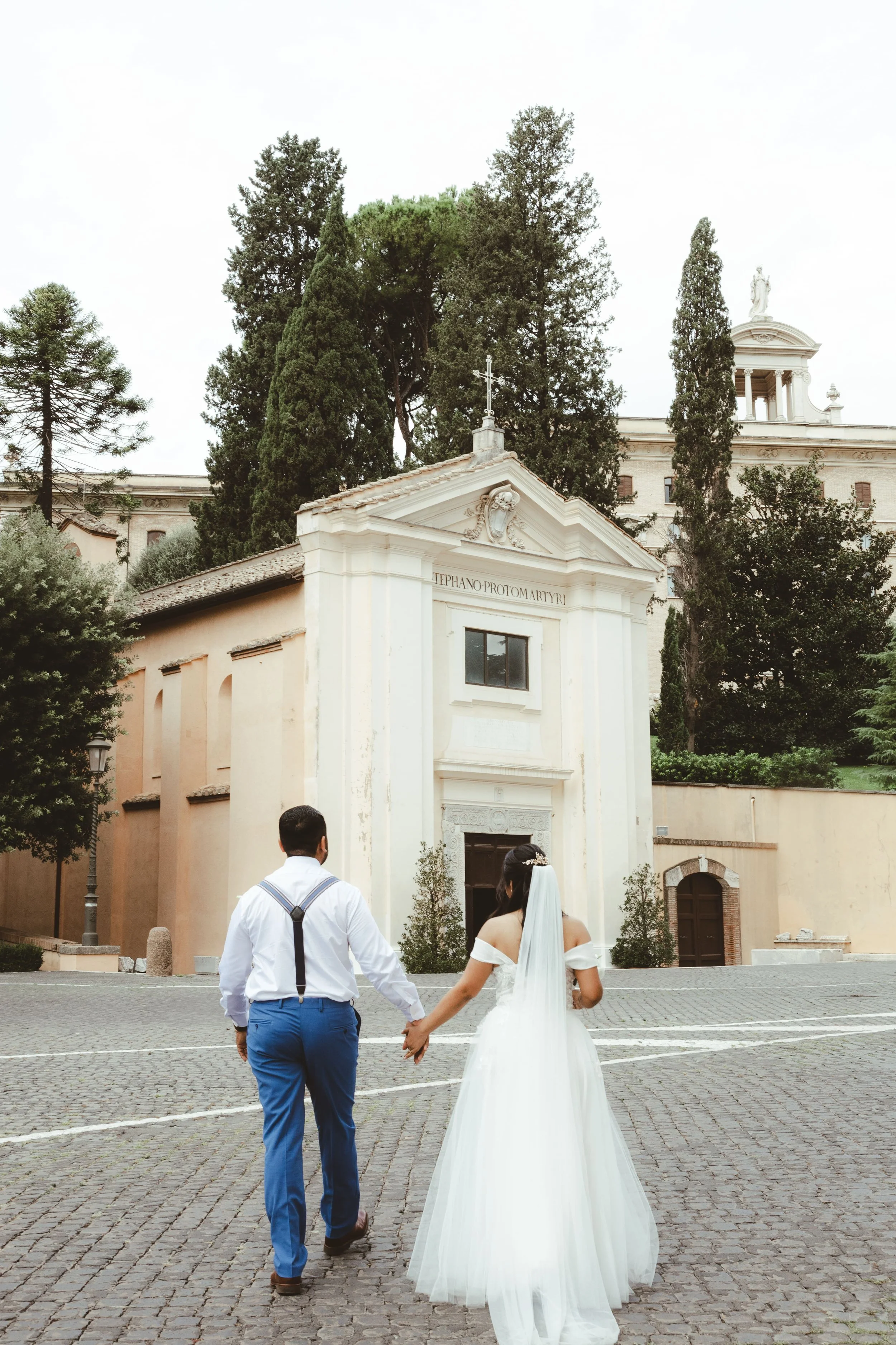 A bride and groom walk hand in hand outside a church, with the church and tall trees in the background.