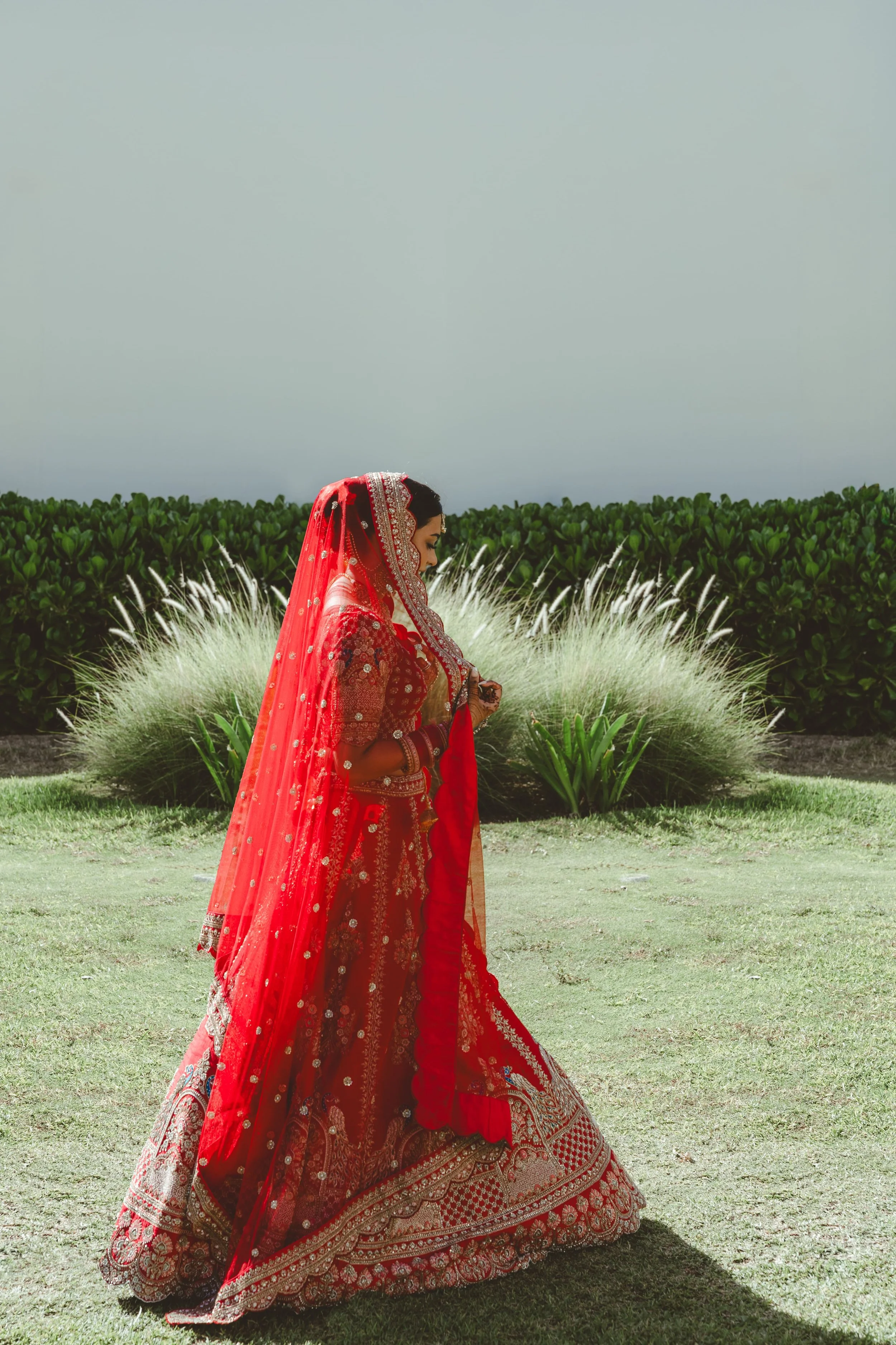 A woman dressed in a red traditional Indian outfit with intricate embroidery, standing outdoors on a grassy area with green bushes in the background, under a light gray sky.