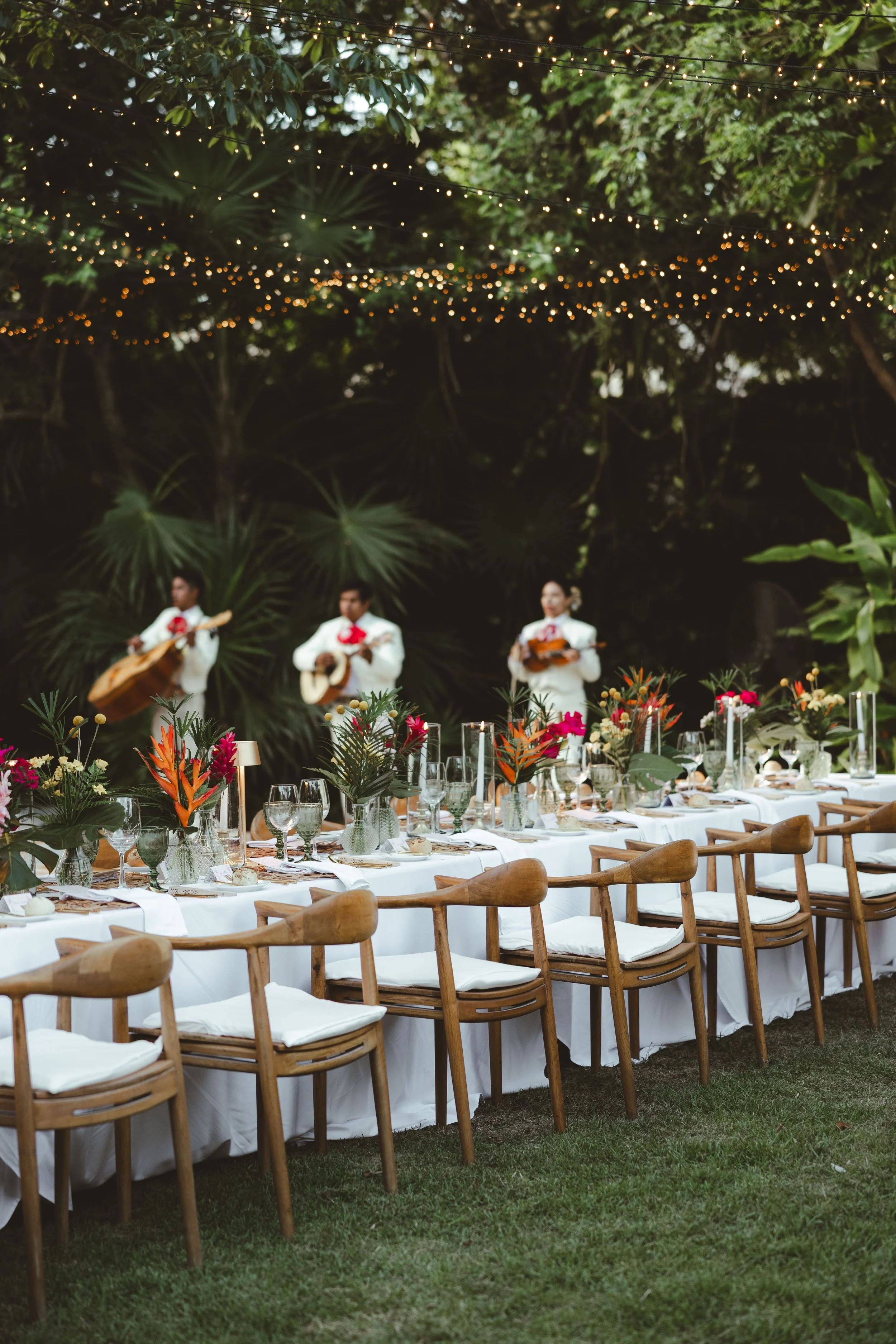 Outdoor wedding reception setup with a long table decorated with flowers, surrounded by wooden chairs, under string lights, with three musicians playing guitar in the background.