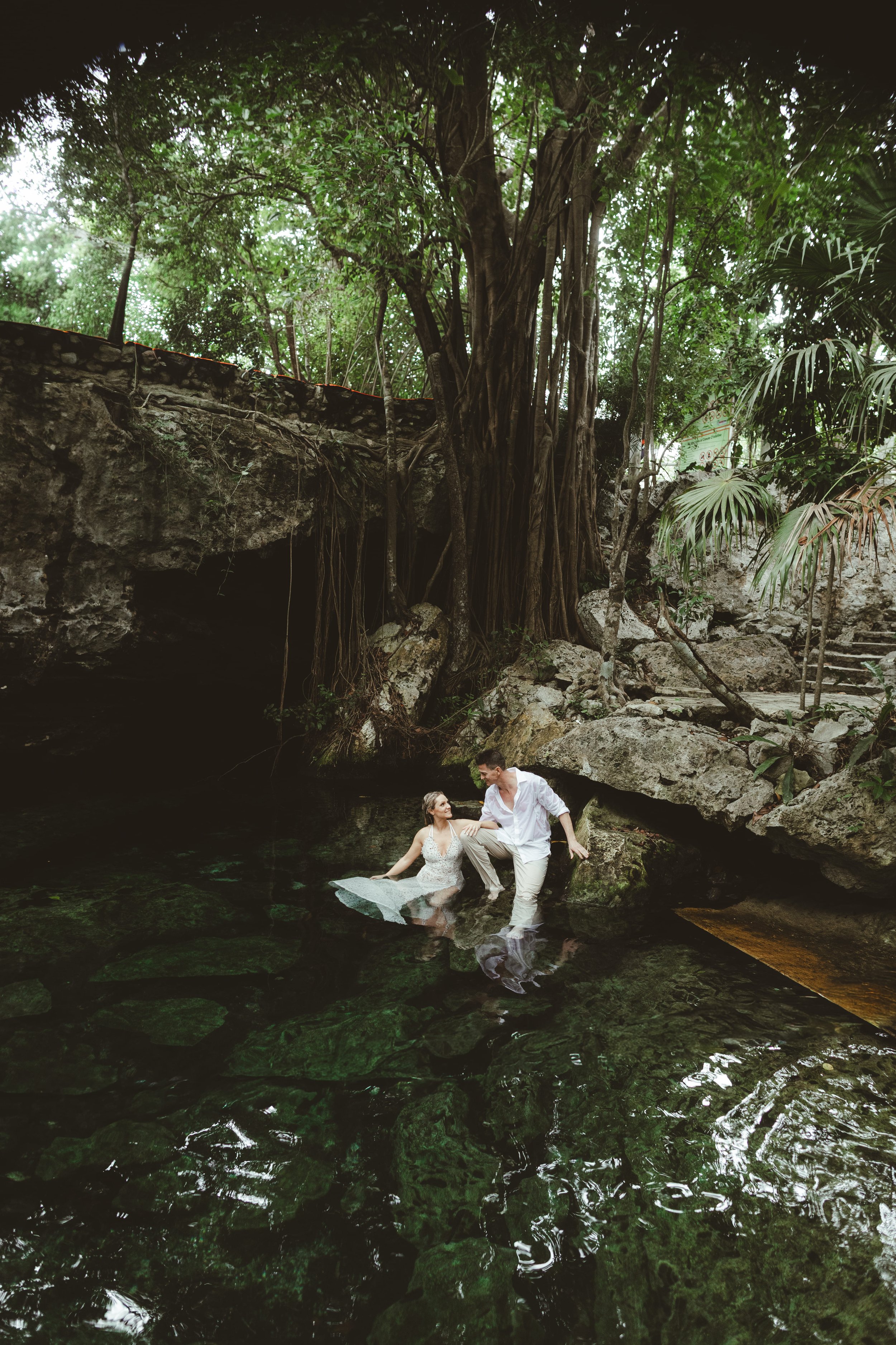 A couple dressed in wedding attire is in a natural pond surrounded by rocks and lush greenery, with tall trees overhead. The woman is sitting on the edge of the water, and the man is standing next to her, both smiling and looking at each other.