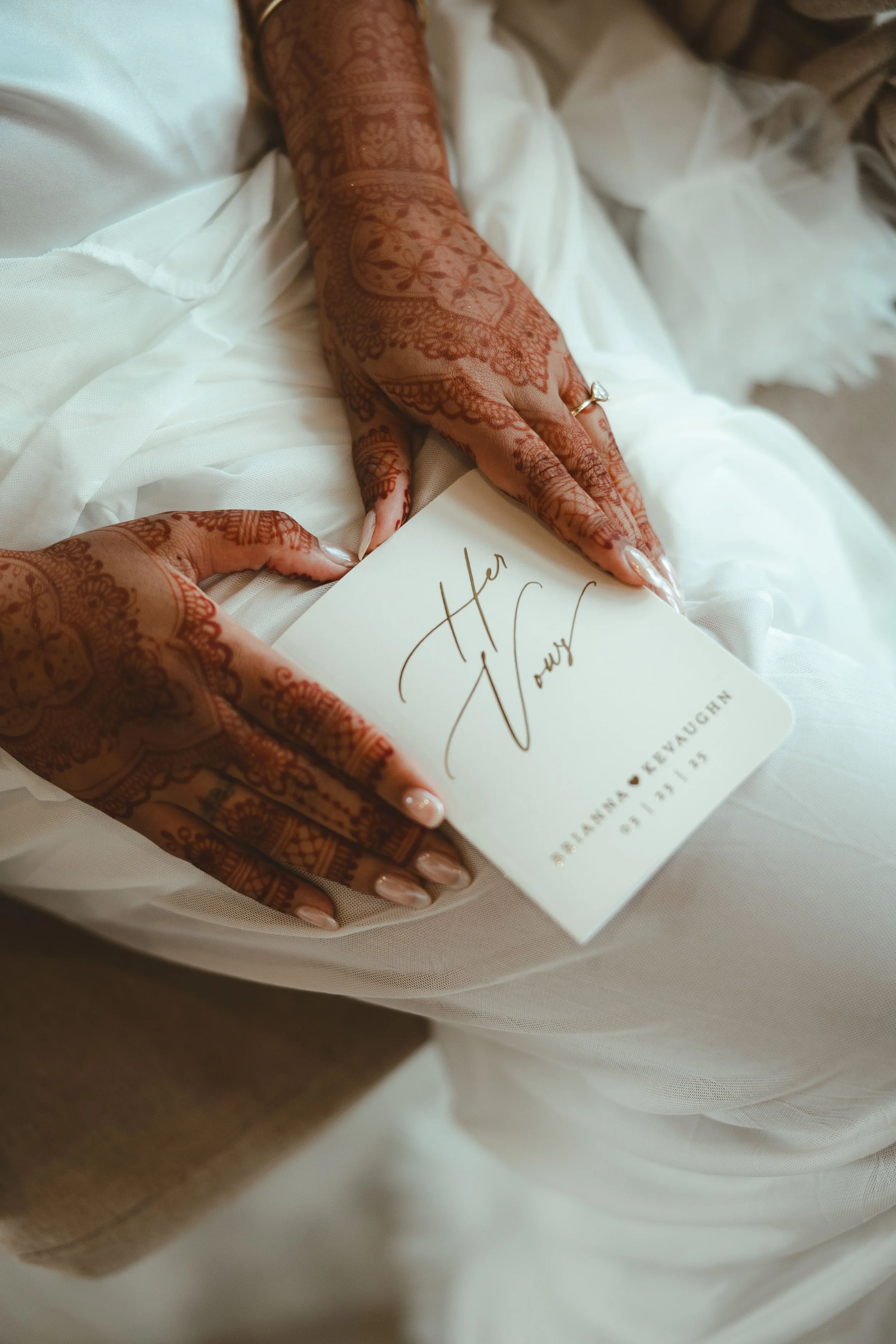 Hands with intricate henna designs holding a white card with elegant handwriting. The card has a name and a date, suggesting a wedding or special event.