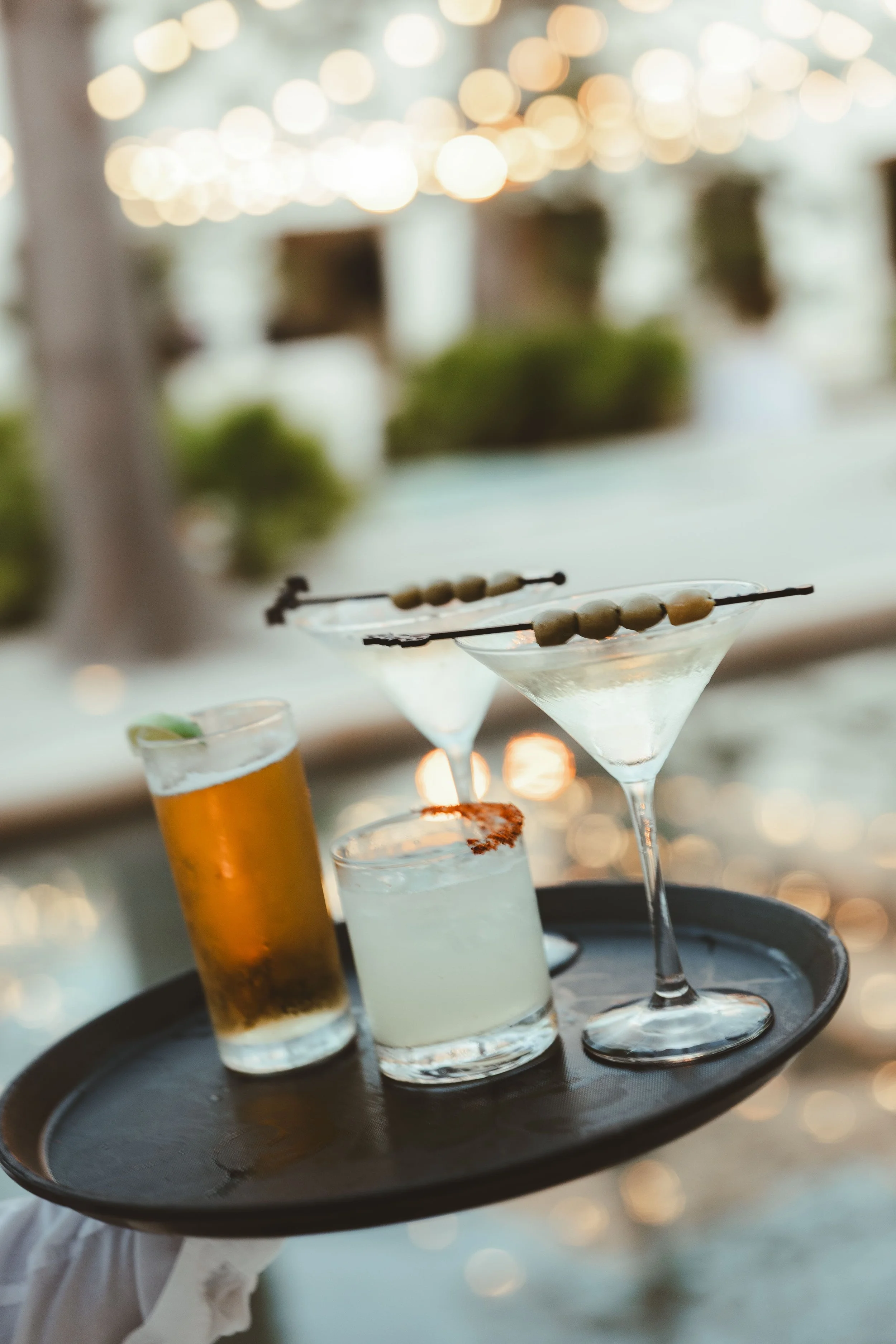 A black tray holds several cocktails with blurred warm lights in the background. The drinks include martinis with olive garnishes, a cocktail with a slice of lime, and a drink with grilled shrimp on the rim.