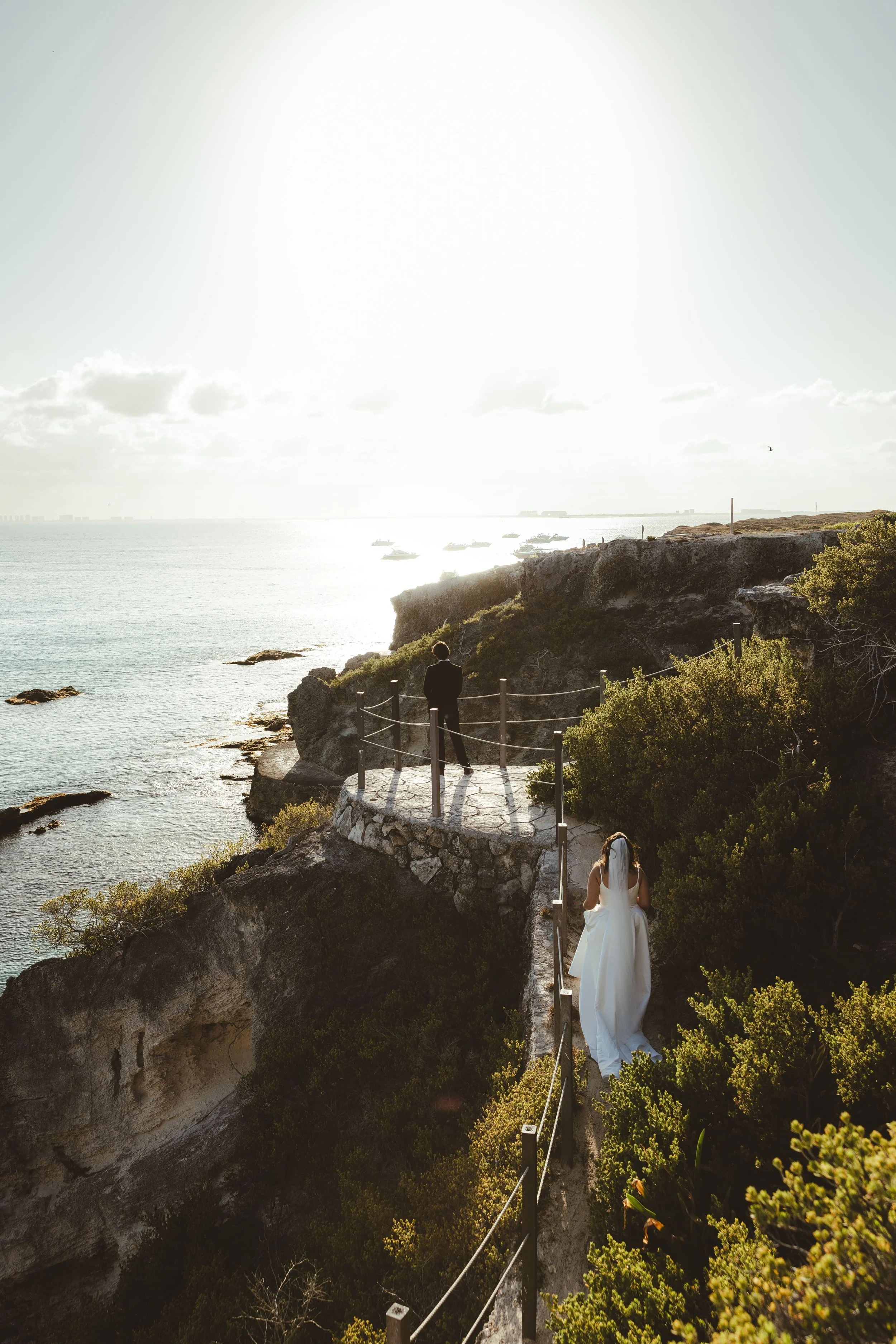 A bride in a white wedding dress walking down a rocky coastal path with a railing, overlooking the ocean with boats and a distant city skyline, during sunset or sunrise.