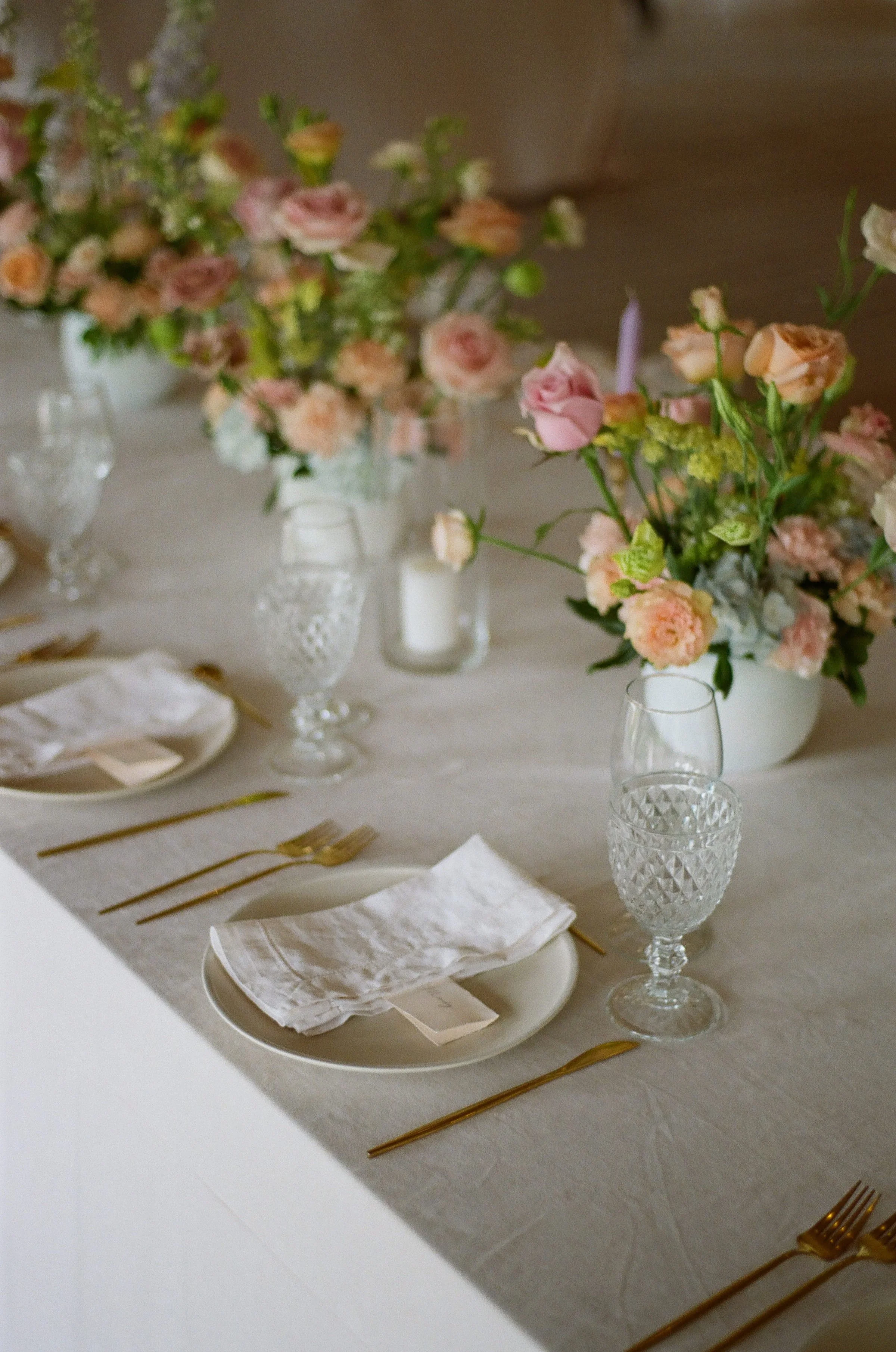 Elegant table setting with clear crystal glasses, gold flatware, white napkins, and plates, featuring pink and peach flower centerpieces in vases.