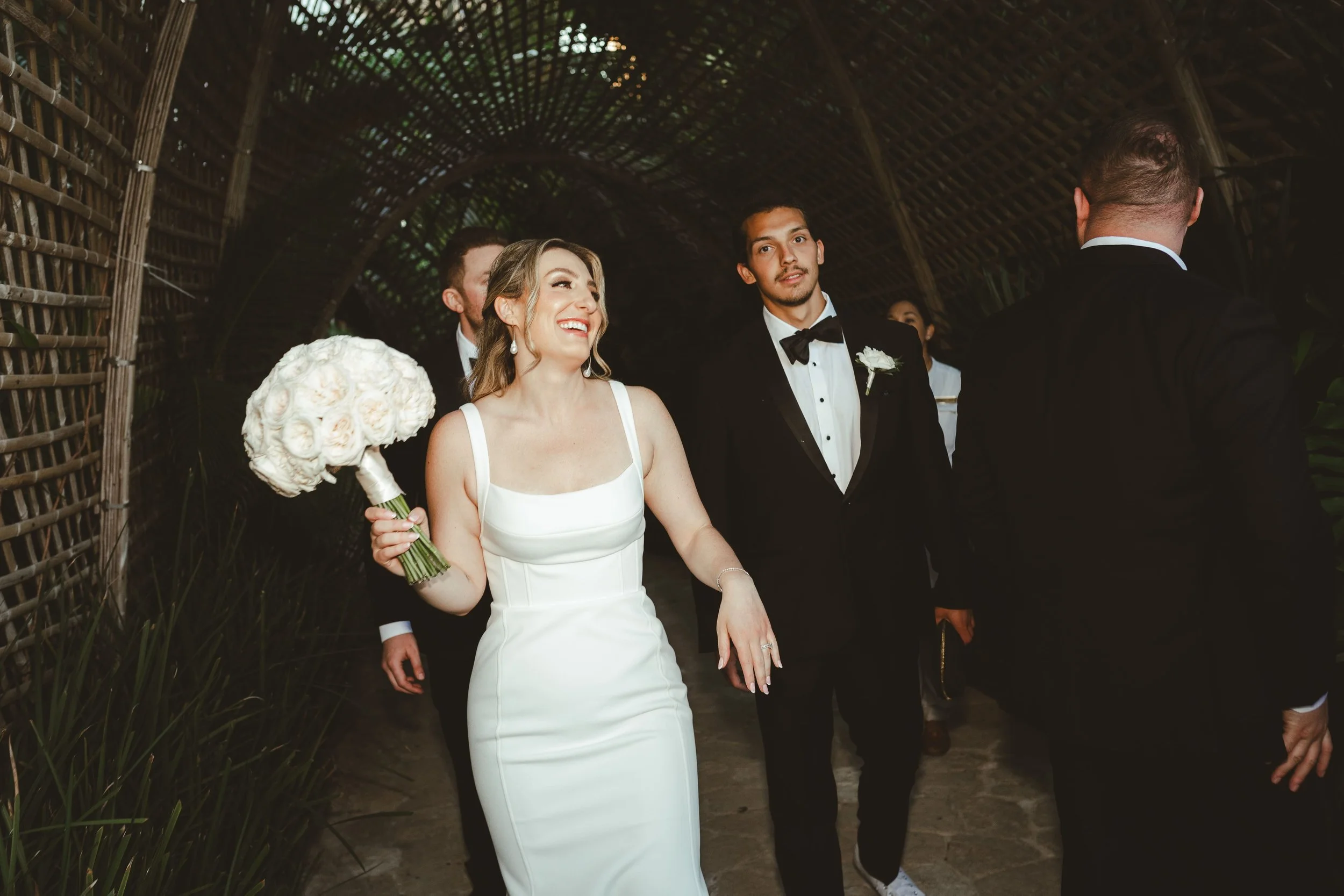 A bride in a white wedding gown holding a bouquet of white roses walking with her groom in a black tuxedo at a wedding ceremony. They are under a woven, bamboo tunnel structure with a lush green background.