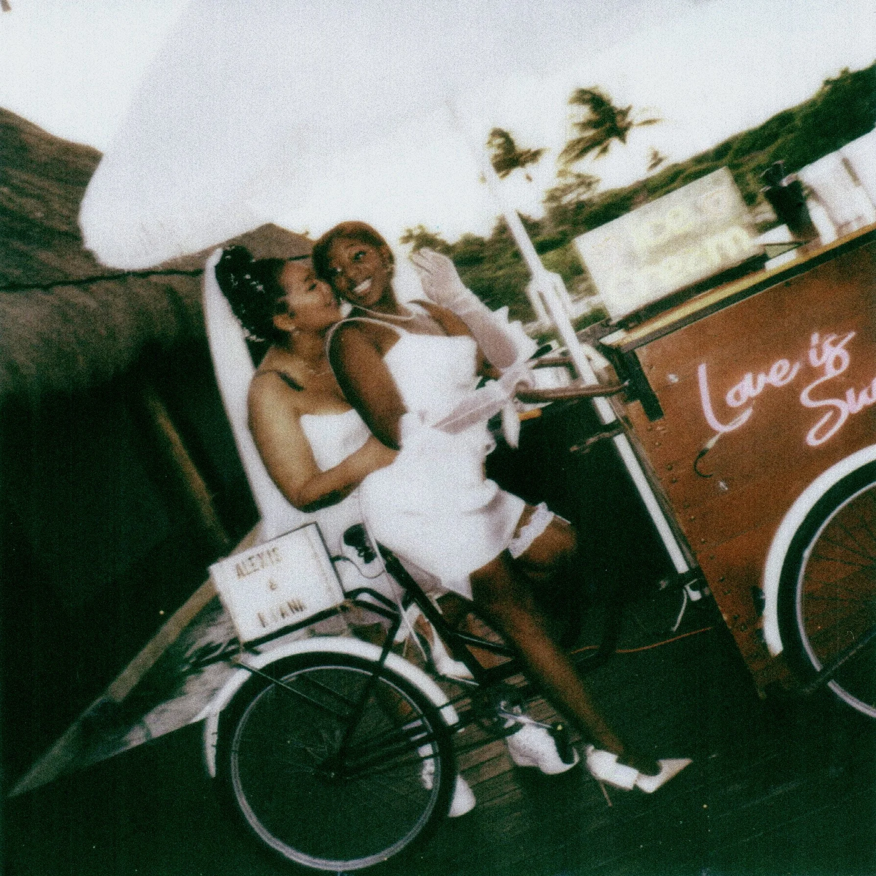 Two women in white dresses lying on lounge chairs outdoors with palm trees in the background, smiling and enjoying the sunny weather at a poolside or beach setting.