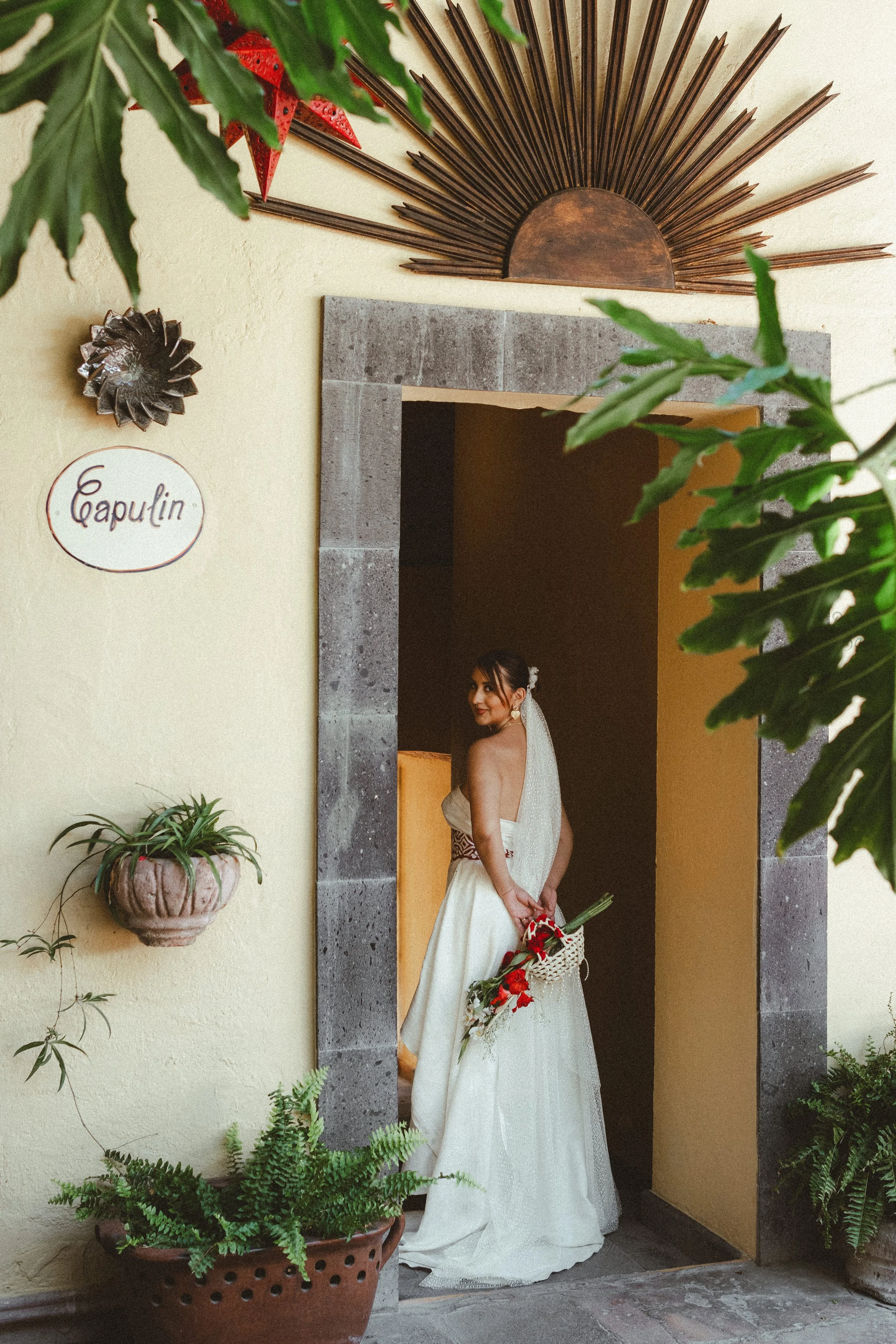 A bride dressed in a white wedding gown with a veil and floral hair accessory standing in a doorway, holding a bouquet of red and white flowers with greenery, surrounded by potted plants and decorative wall hangings.
